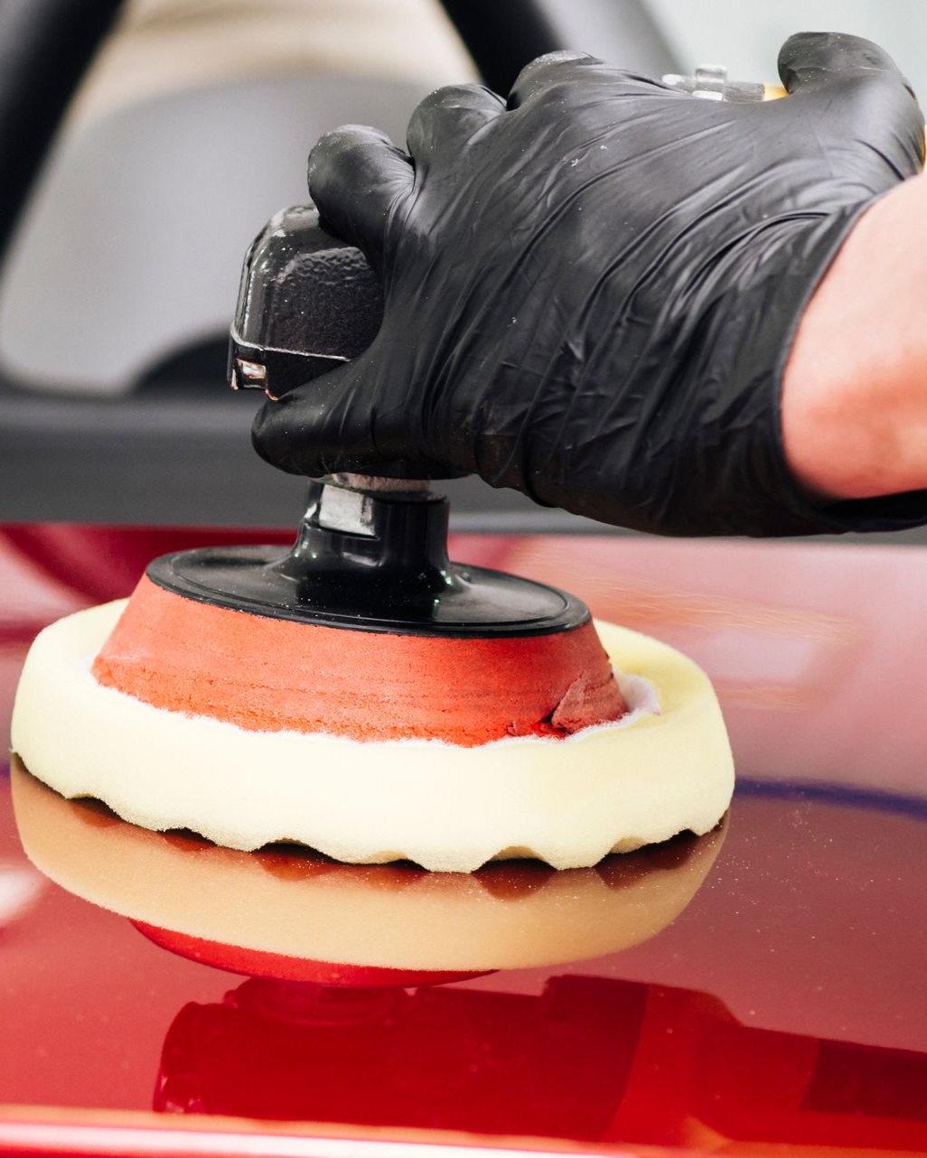 A hand wearing a black glove uses a red and white foam buffing pad to polish a shiny red car surface.