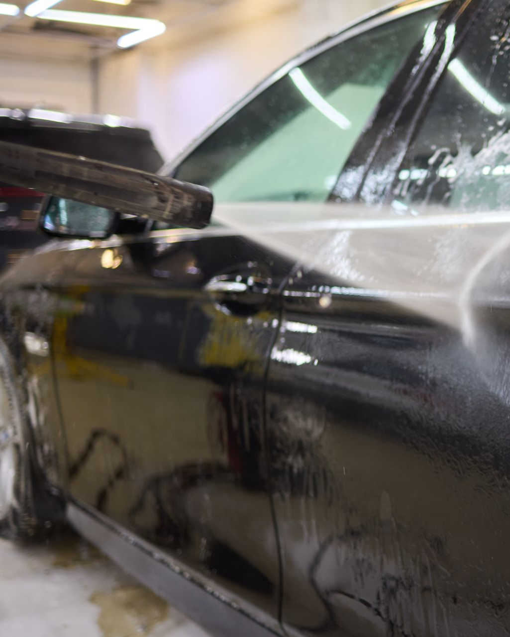 A close-up of a pressure washer spraying water onto the wet, black door of a car in a professional garage.