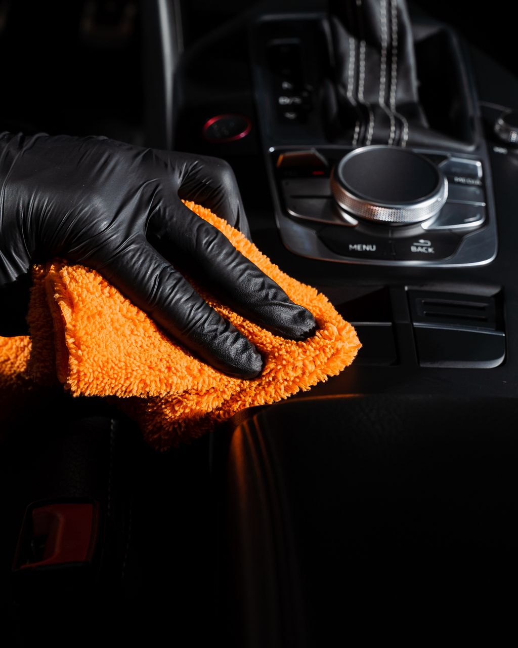 A hand in a black glove uses an orange microfiber cloth to clean a car's black center console.