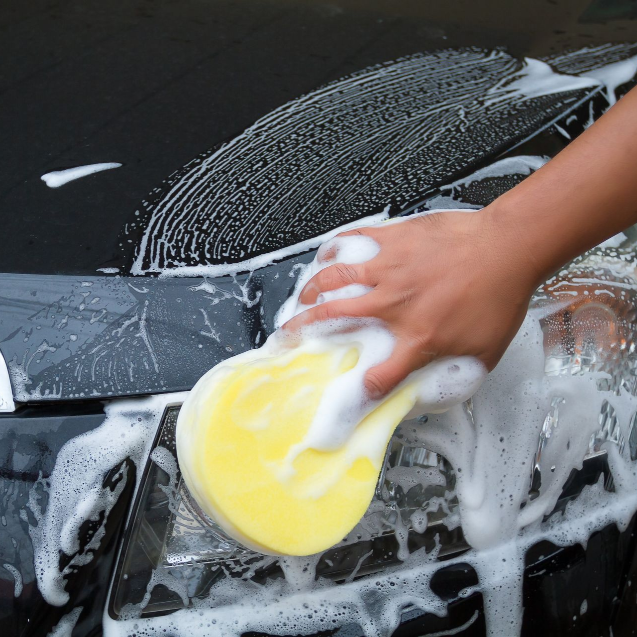 A hand uses a yellow sponge to wash soap suds off the headlight of a black car.