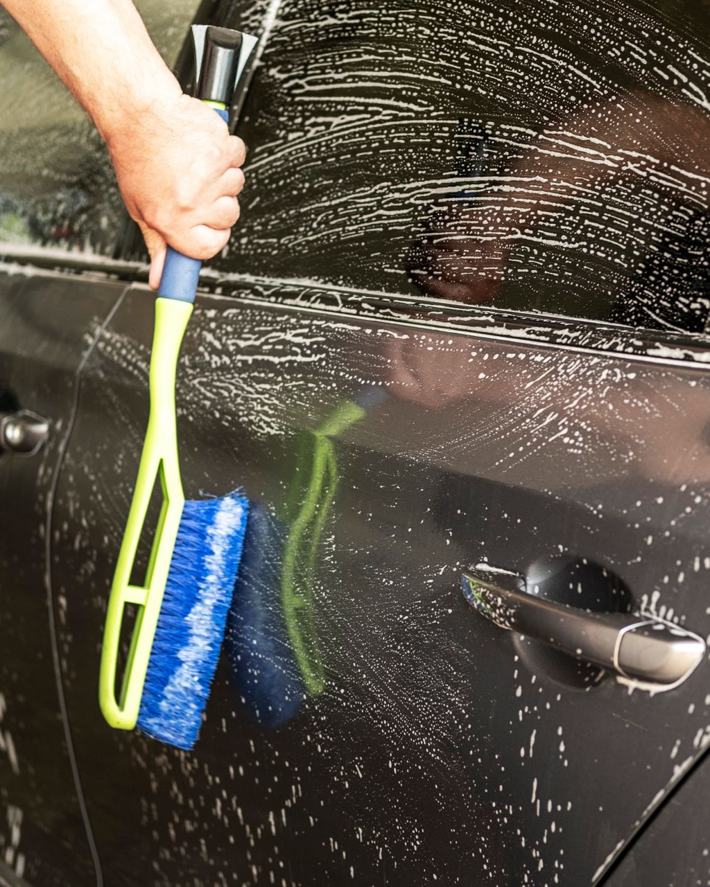 A person uses a lime-green long-handled brush with blue bristles to wash a sudsy black car door.