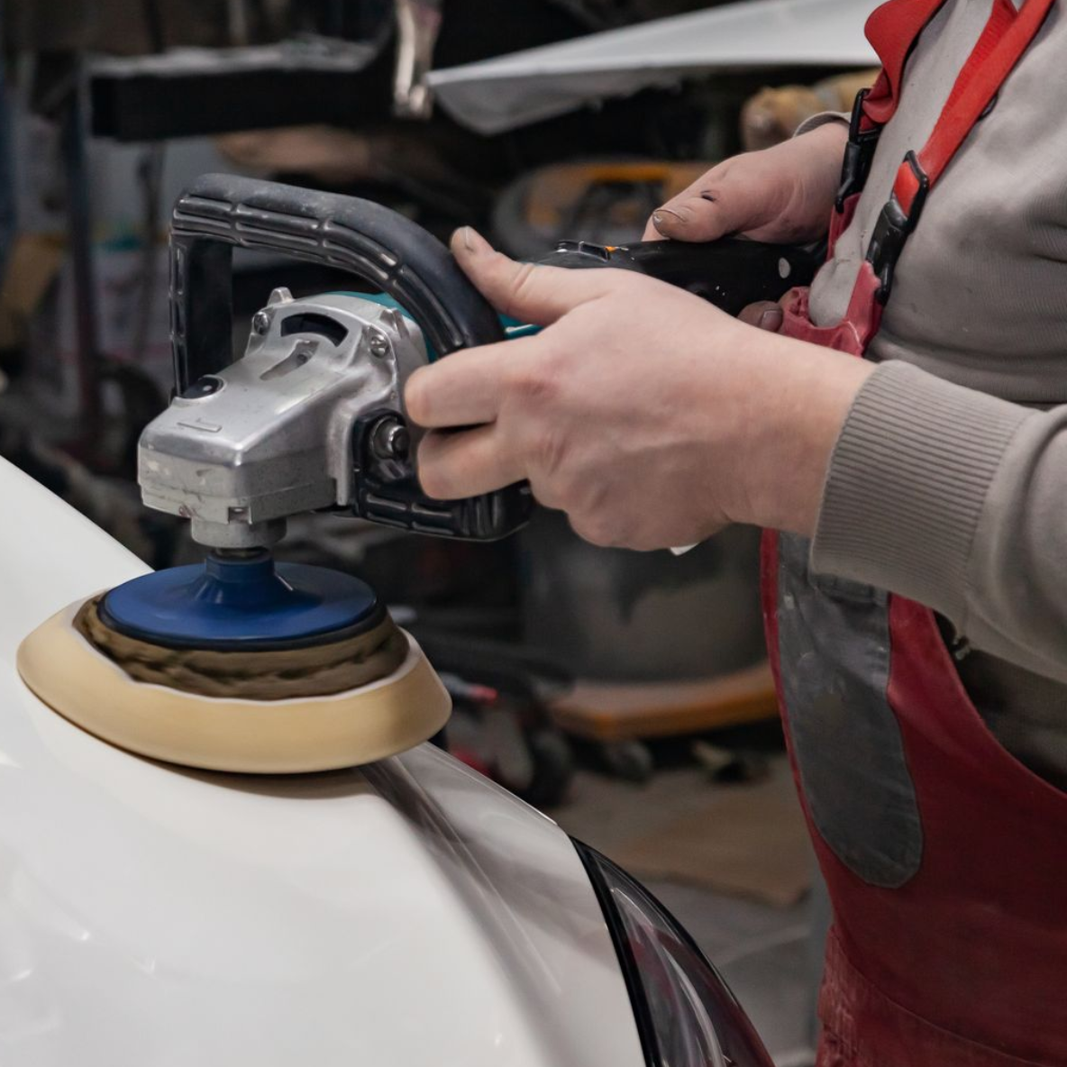 A person wearing red overalls uses an electric buffer to polish the white exterior of a vehicle in a workshop.