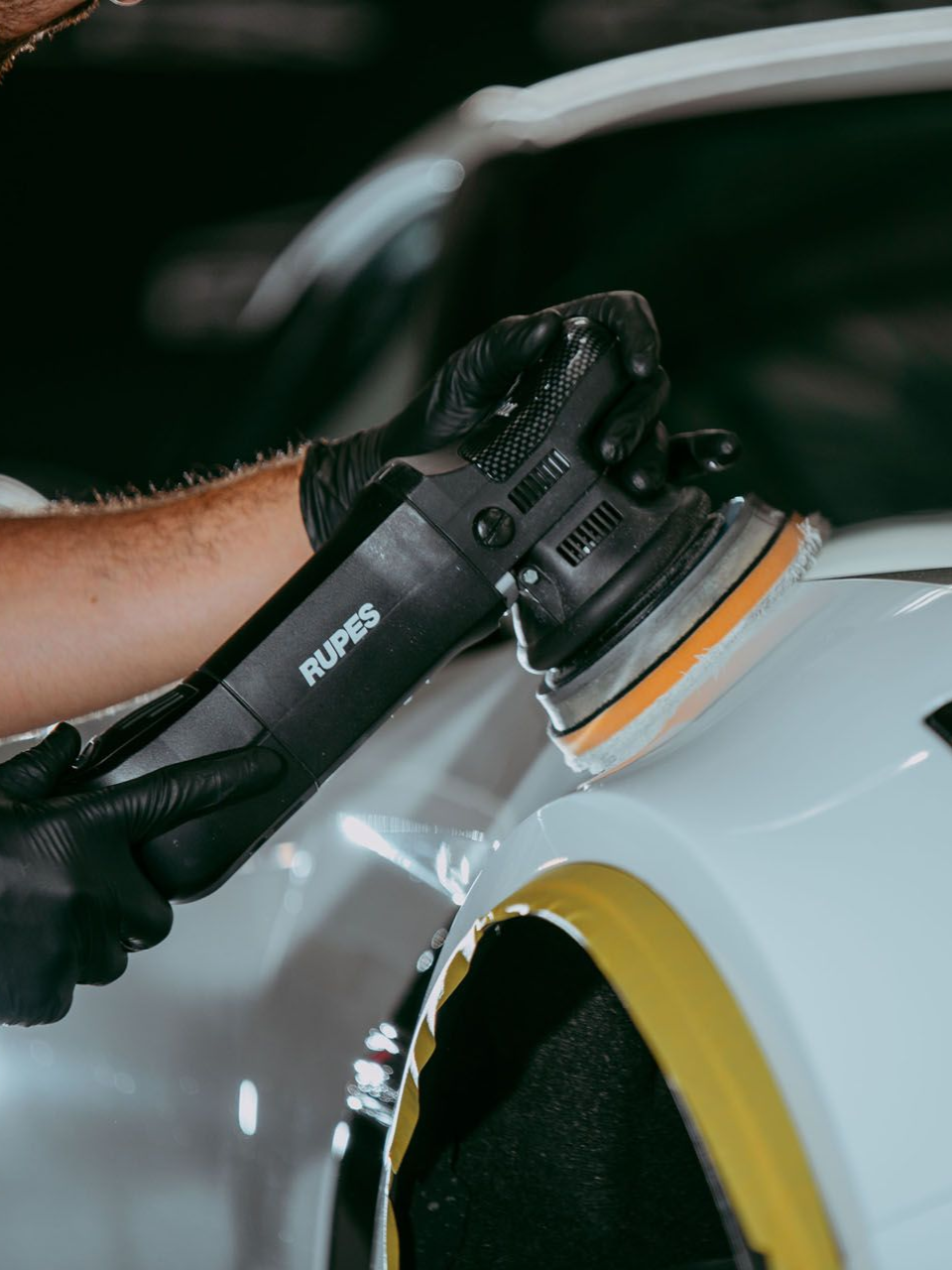 A person uses a handheld power polisher to buff the white paint on a vehicle.