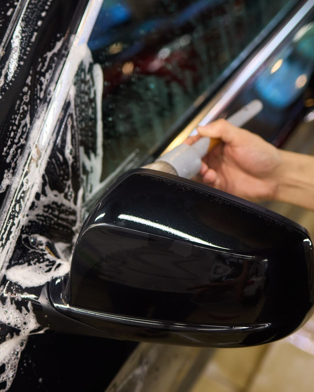 A hand uses a soft brush to clean soap suds from the black side mirror and door of a car.