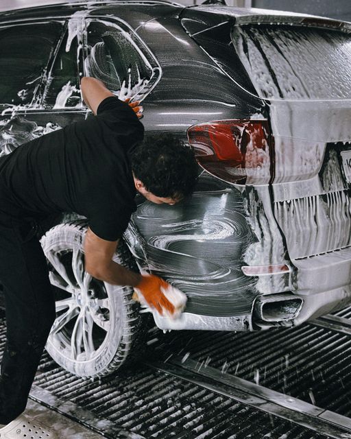 A person in black clothing scrubs a soapy, dark-colored car in a wash bay with an orange sponge.