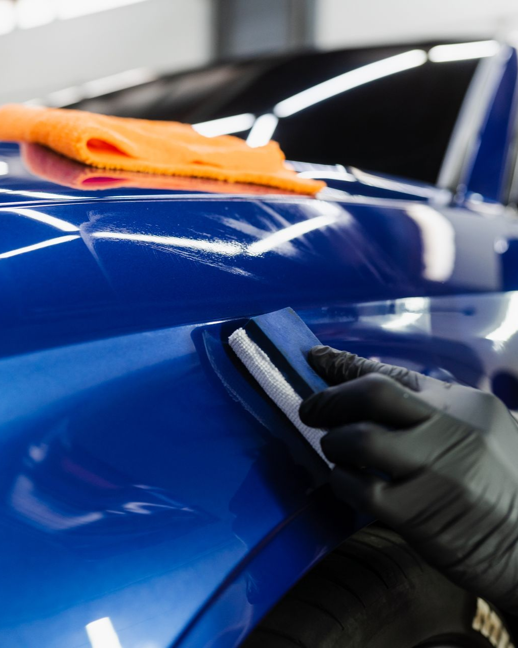A gloved hand uses a foam applicator block to apply a ceramic coating to a glossy blue car panel.