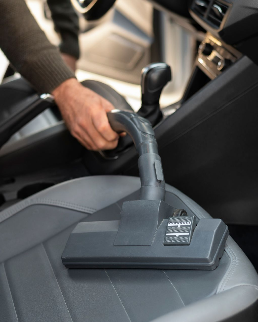 A person uses a vacuum cleaner attachment to clean the gray upholstery of a car seat.