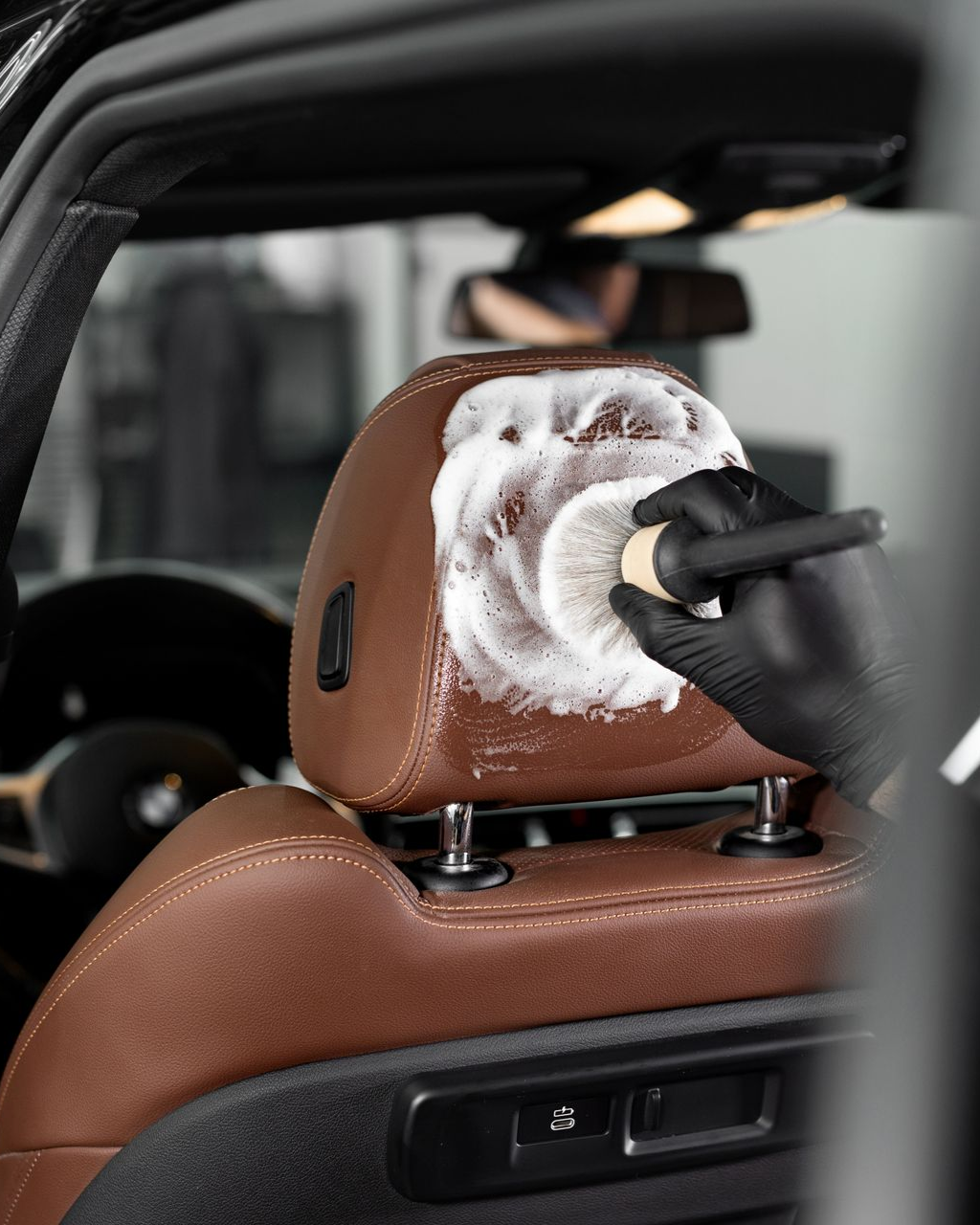 A gloved hand uses a brush and foam cleaner to scrub a brown leather car headrest.