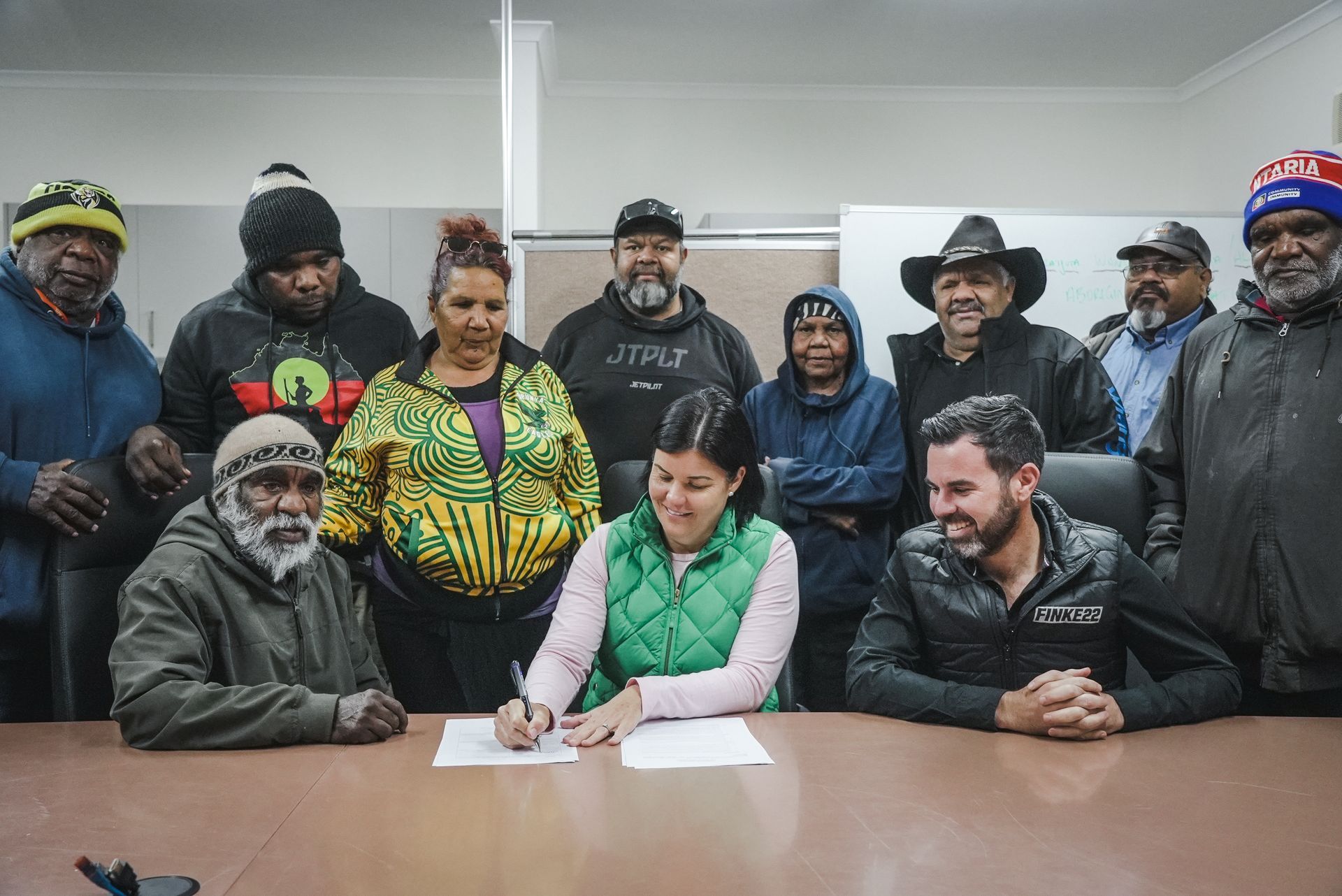 Group of people, including a woman signing a document, at a table. Men in hats and jackets stand behind them.