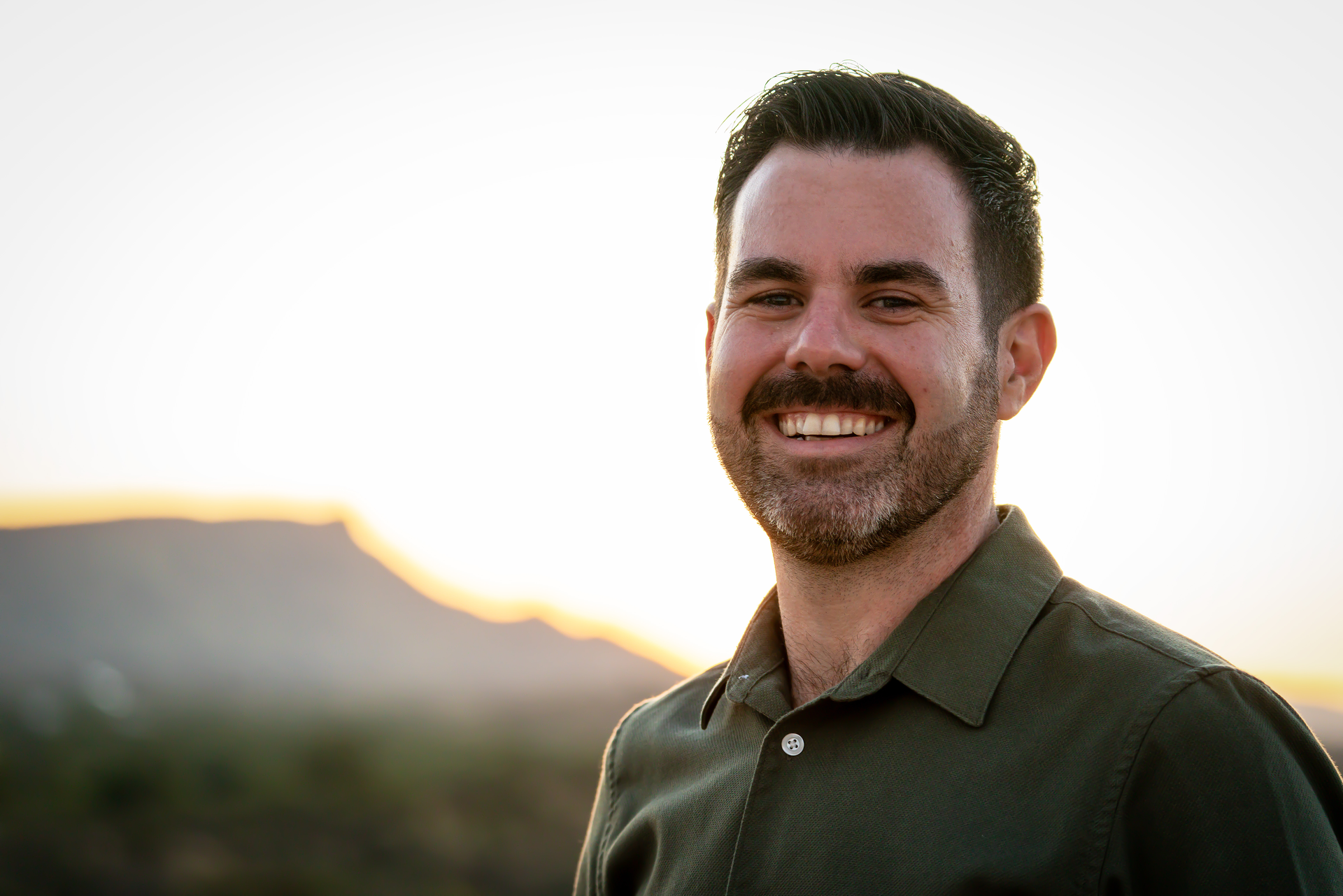 Man with dark hair and beard smiles broadly outdoors against a sunset backdrop.