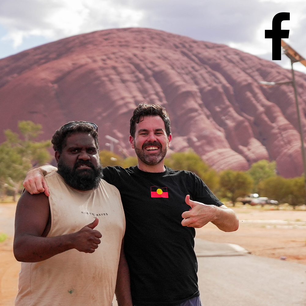Two men with thumbs up in front of Uluru, Australia. One wears a tank top, the other a black shirt with an Aboriginal flag.