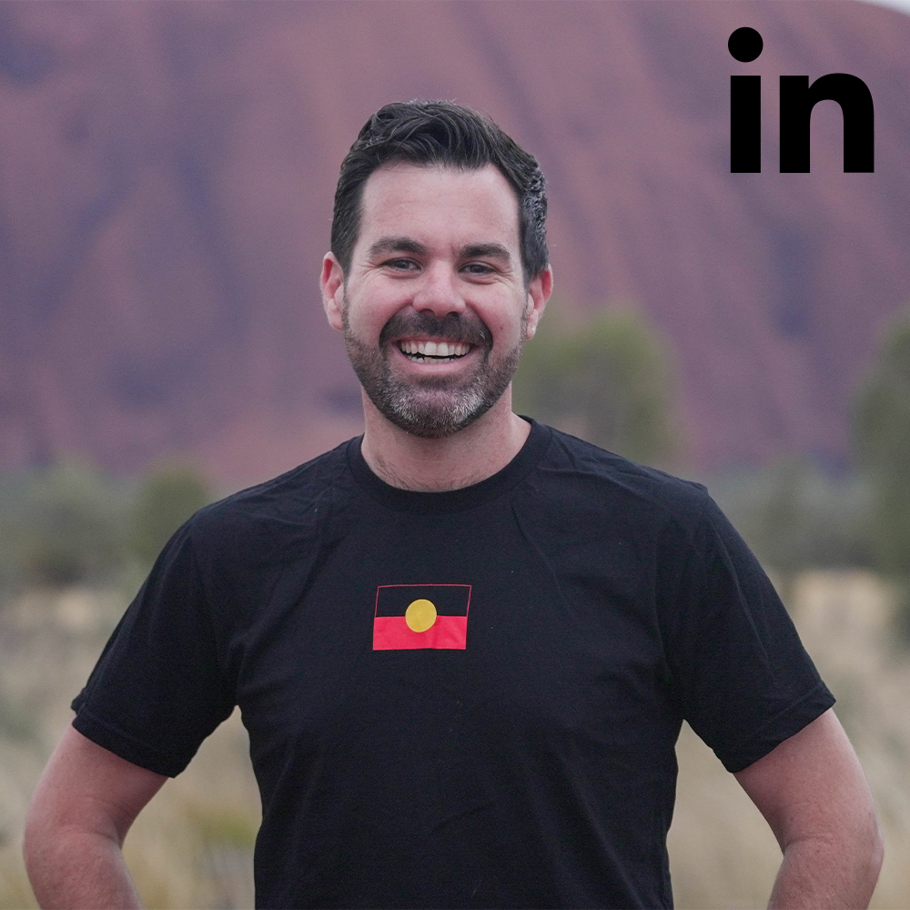 Man smiling, wearing black shirt with Aboriginal flag, outdoors with red rock background. 