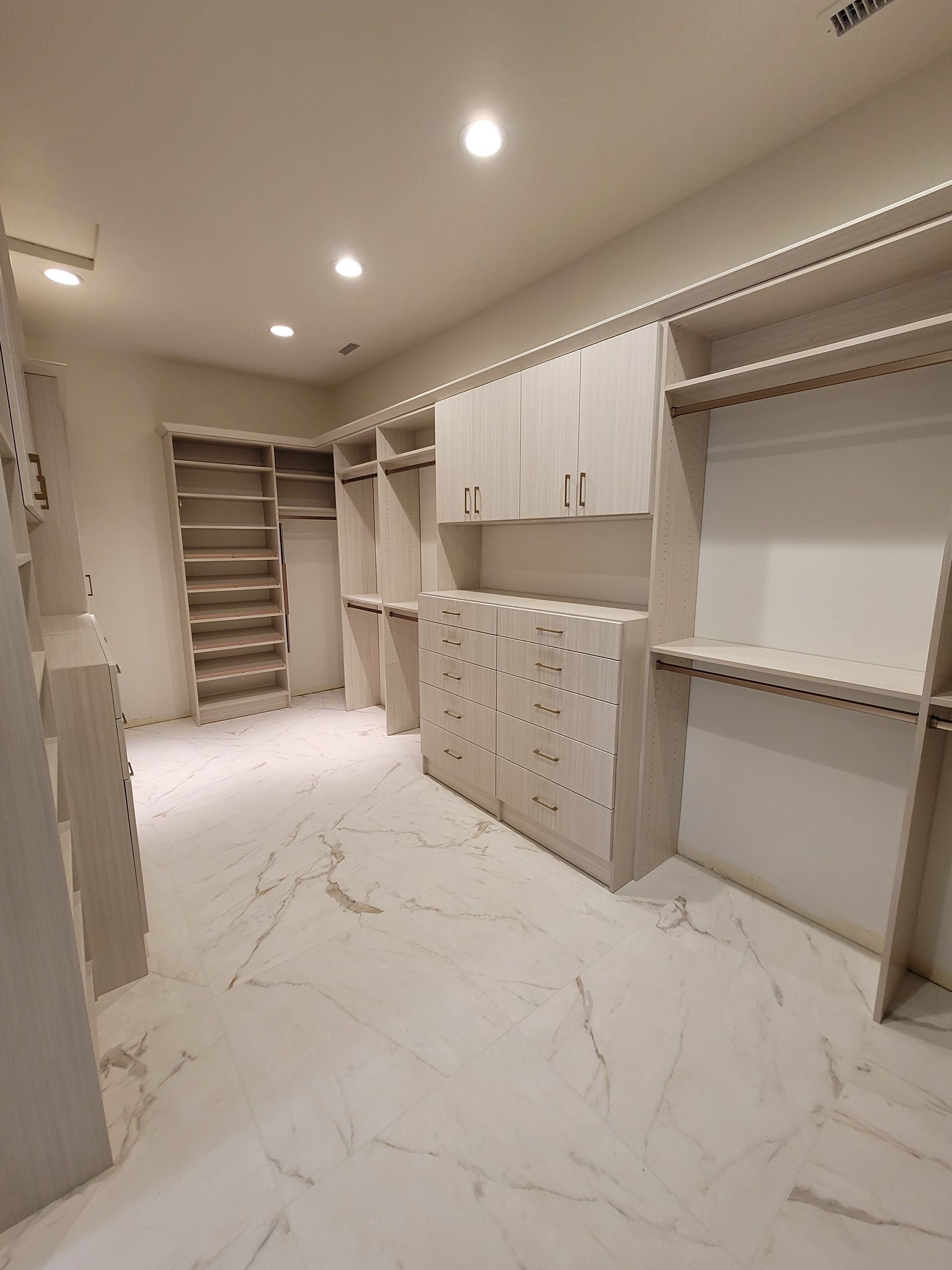 Empty walk-in closet with white built-in shelves, drawers, and marble-look floor under recessed lights