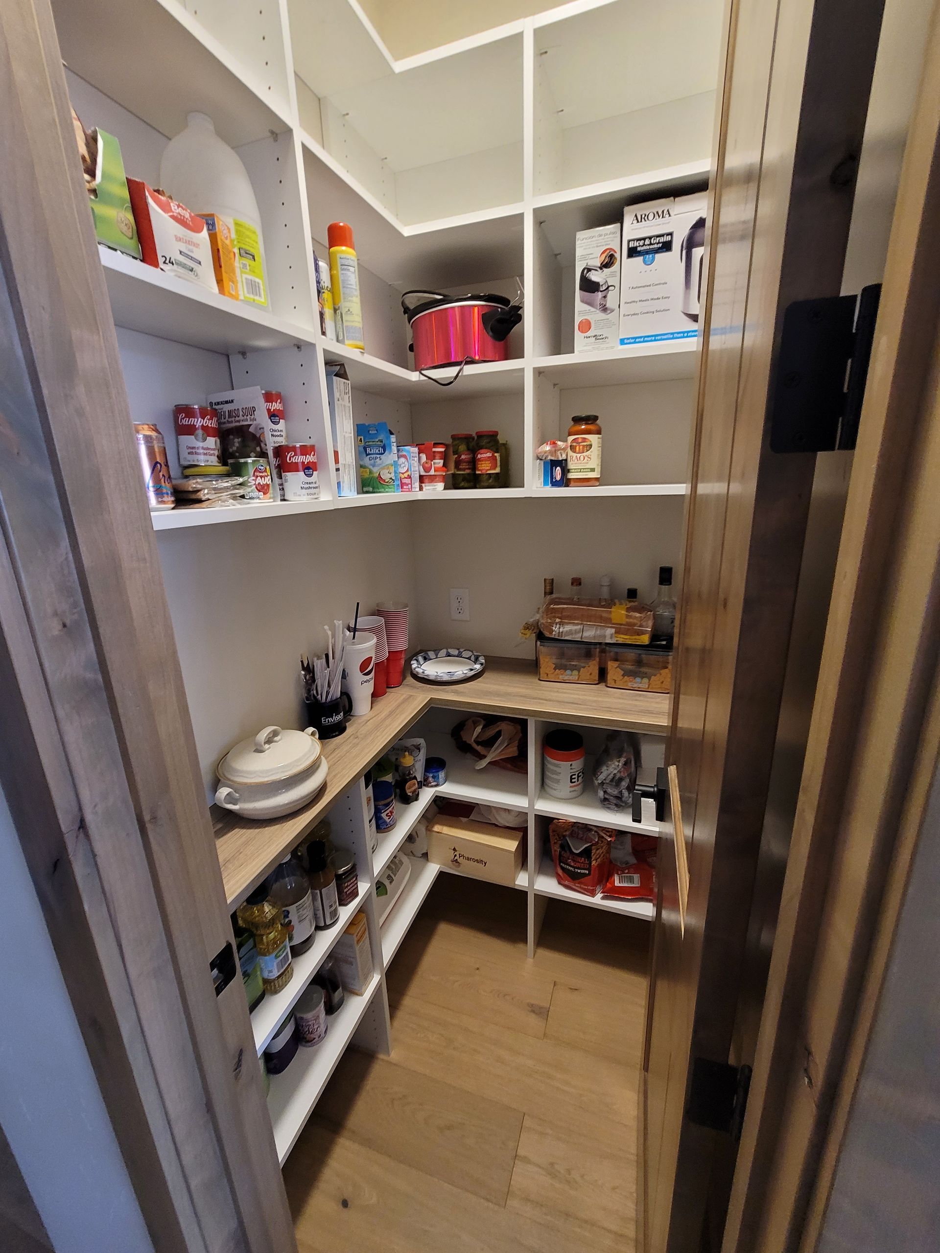Small pantry with white shelves and wooden floor, stocked with food containers, dishes, and household items.