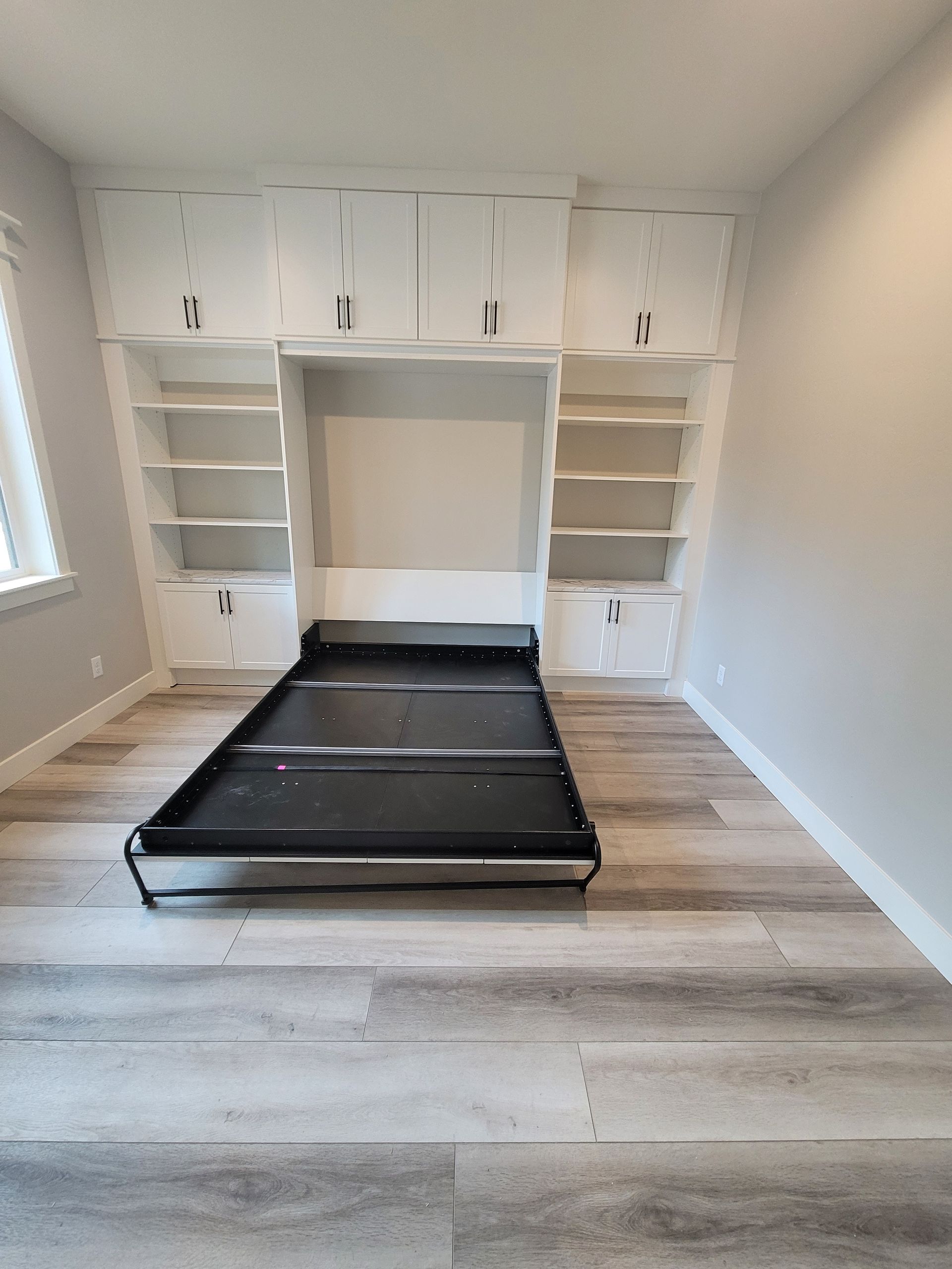Empty bedroom with built-in shelves, white walls, and a black folding bed frame on wood-look flooring