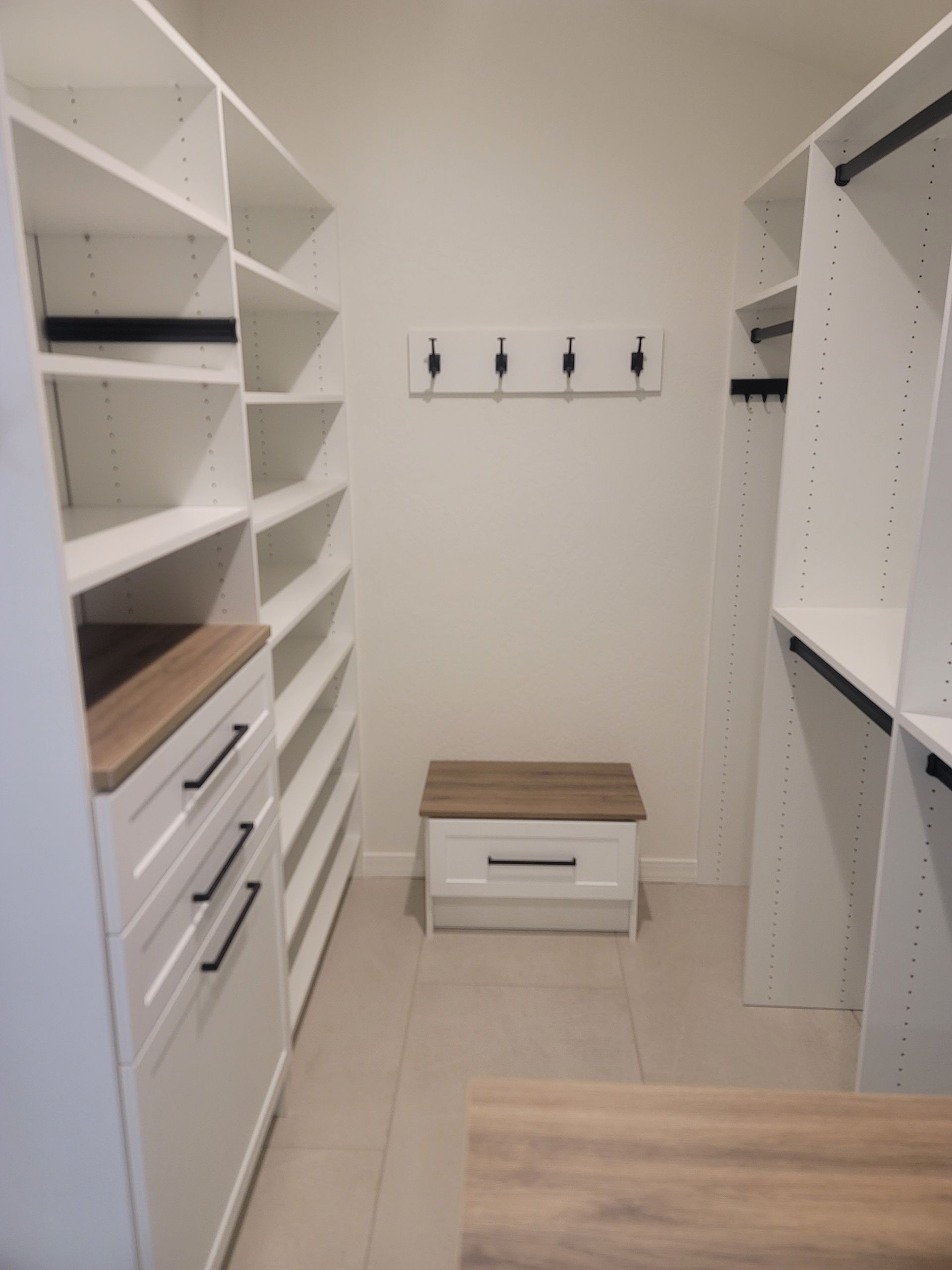 Empty walk-in closet with white shelves, drawers, hooks, and a small bench on beige tile floor.