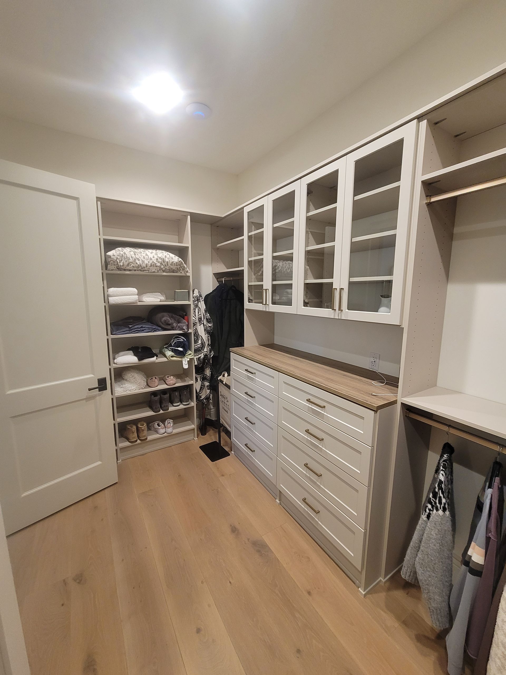 Walk-in closet with white cabinets, drawers, shelves, and hanging clothes on wood flooring.