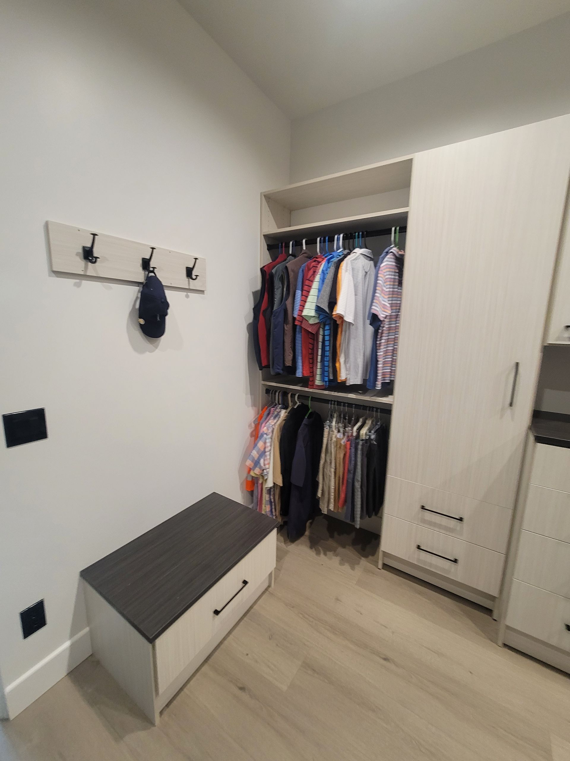 Small walk-in closet with open clothing racks, white cabinets, and a gray bench on light wood flooring.