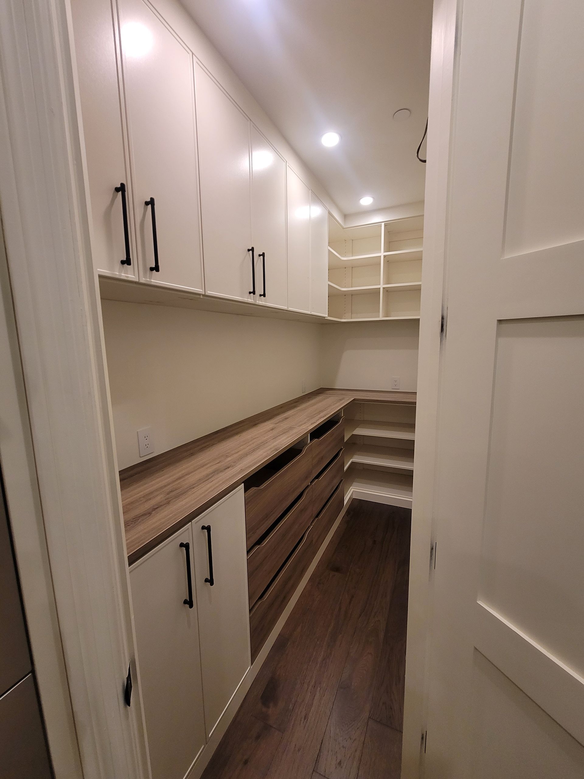 Narrow walk-in closet with white cabinets, long wood countertop, drawers, and built-in shelving under ceiling lights