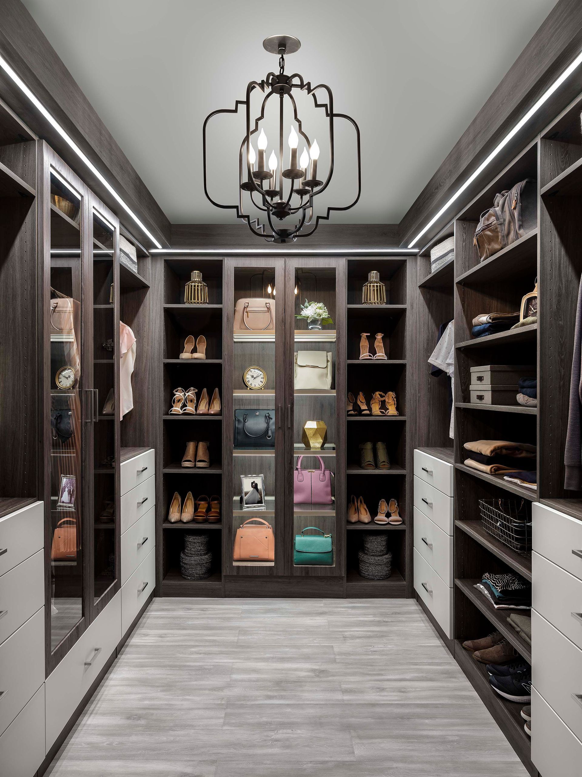 Elegant walk-in closet with dark shelving, glass cabinets, and a chandelier over a light gray floor.