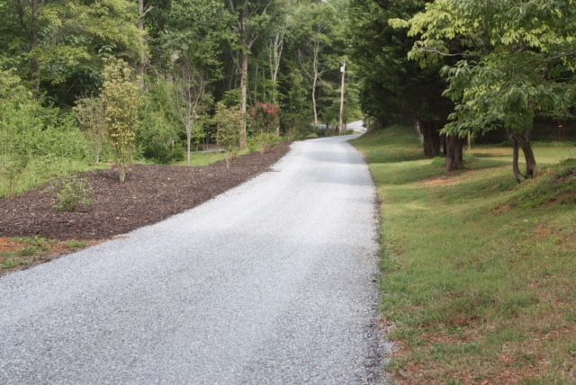 Gravel road winds through a wooded area. One side has grass and trees, the other side has mulch and shrubs.