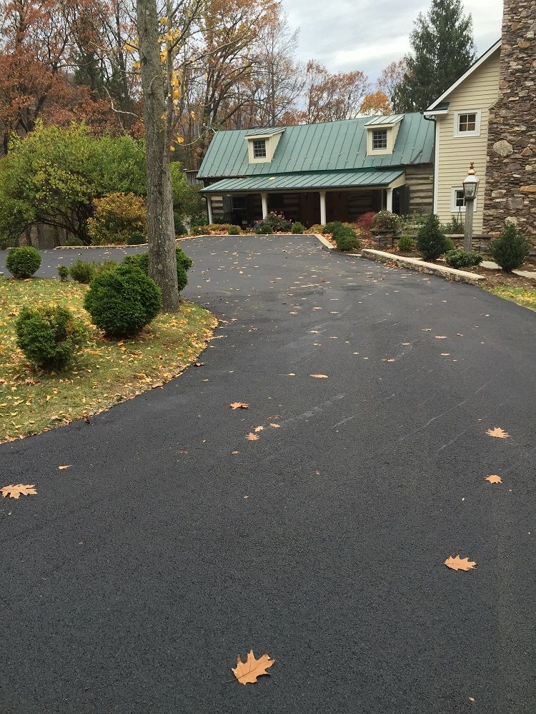 Black asphalt driveway leads to a two-story house with a covered porch and green roof.
