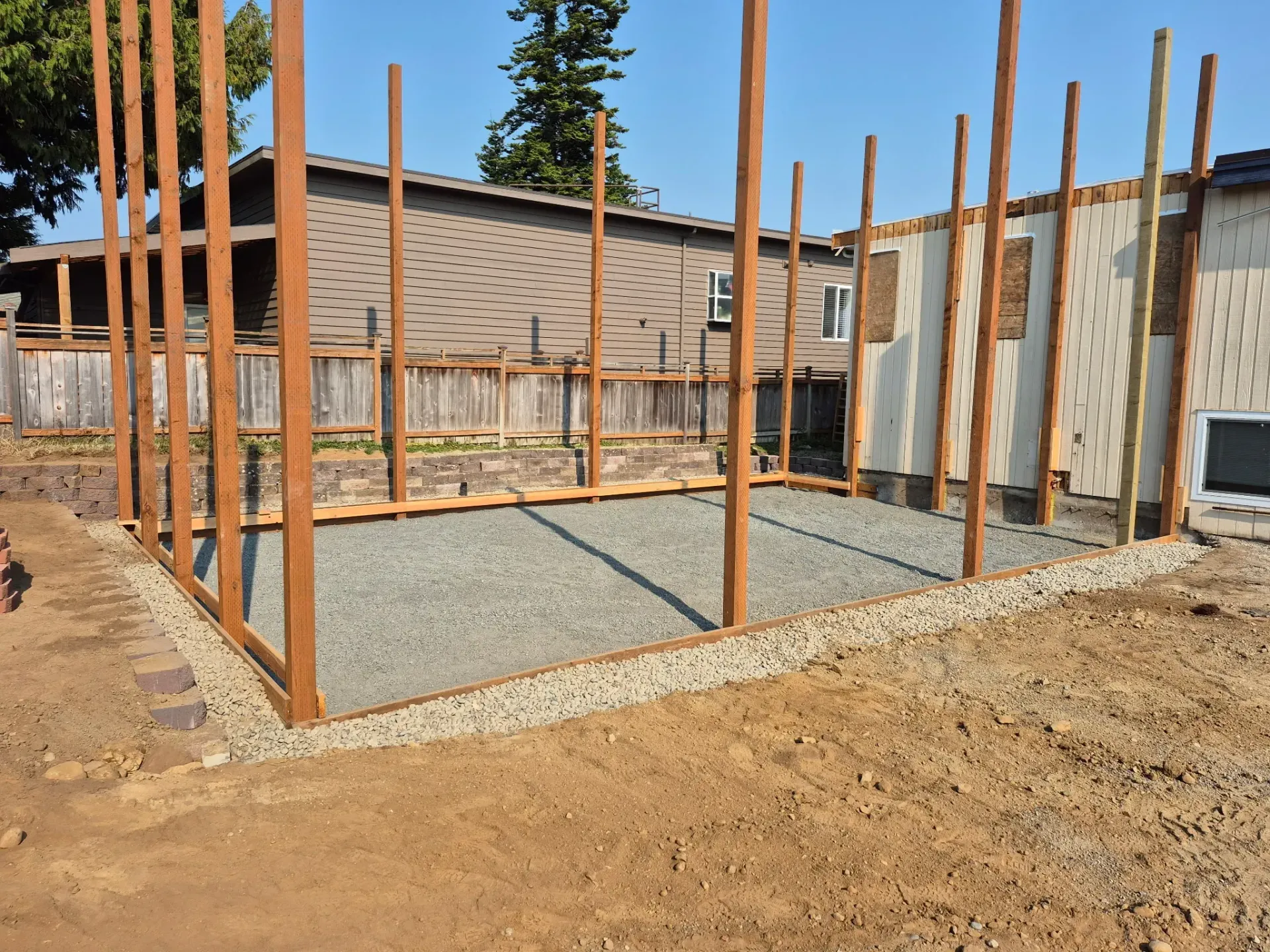 A wooden frame structure under construction on a gravel-filled base in a backyard setting.