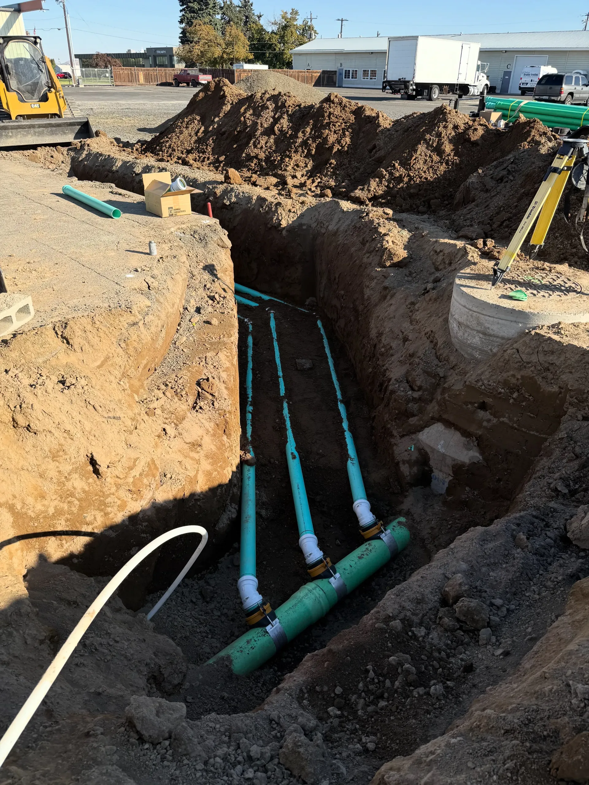 A construction site trench containing a green pipe manifold connected to three smaller blue pipes.