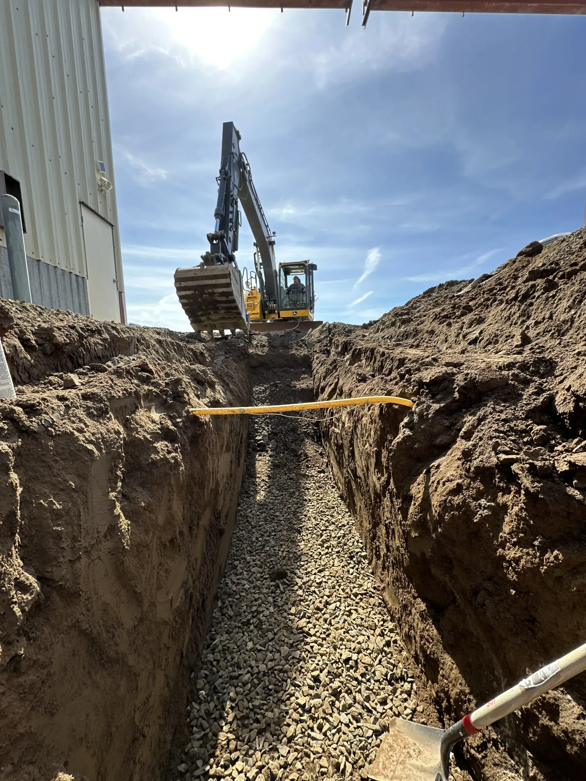 A yellow excavator sits at the far end of a deep dirt trench filled with gravel, viewed from inside the trench.