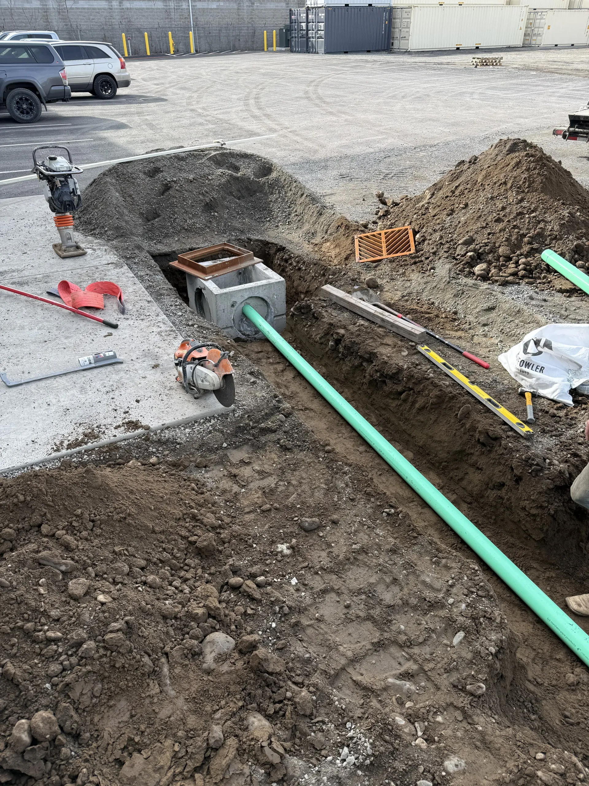 A green pipe runs through a trench next to a concrete utility box, with construction tools scattered on the ground.