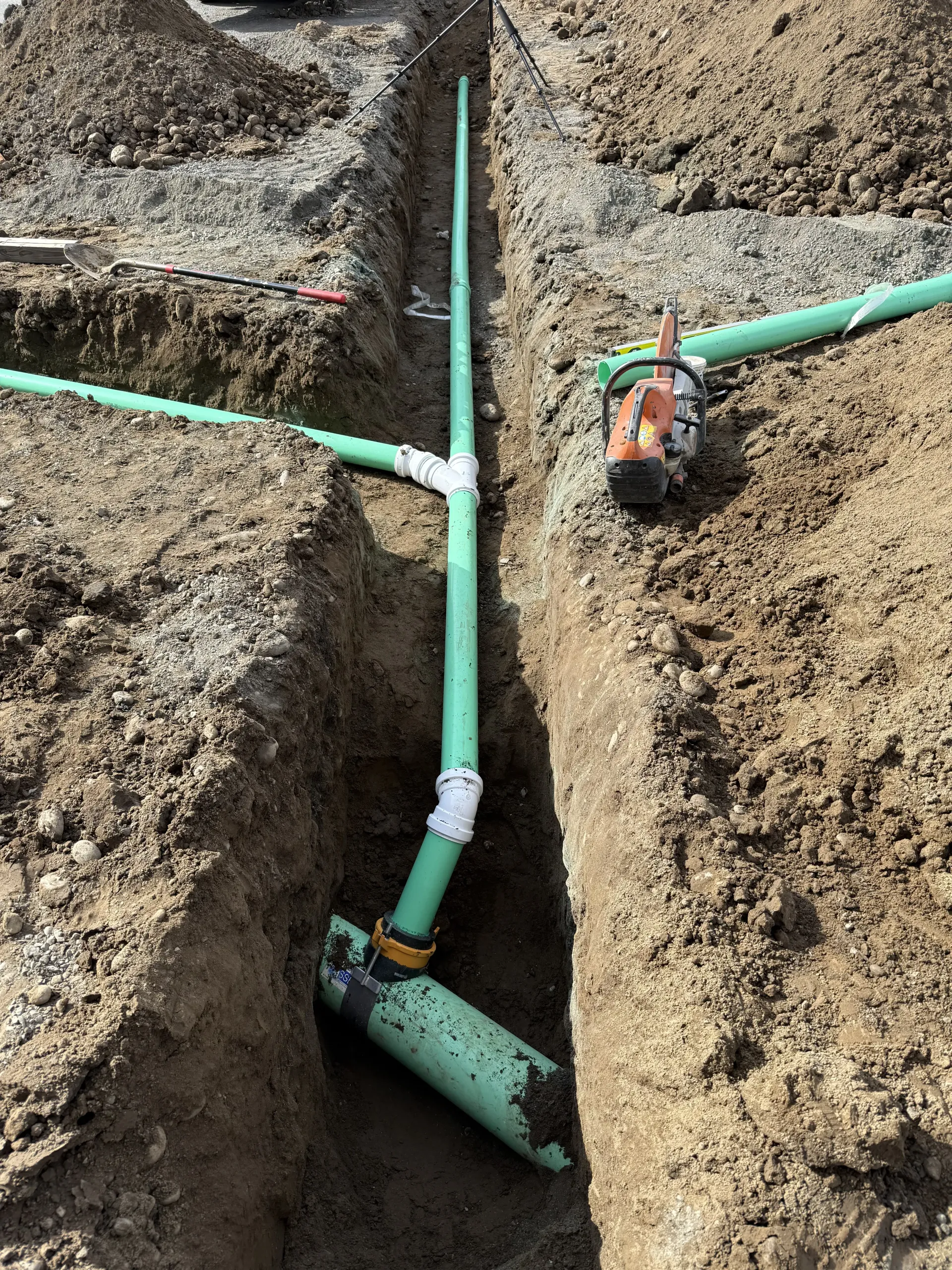 Green PVC pipes with T-junctions and elbows installed in a narrow, dirt construction trench.