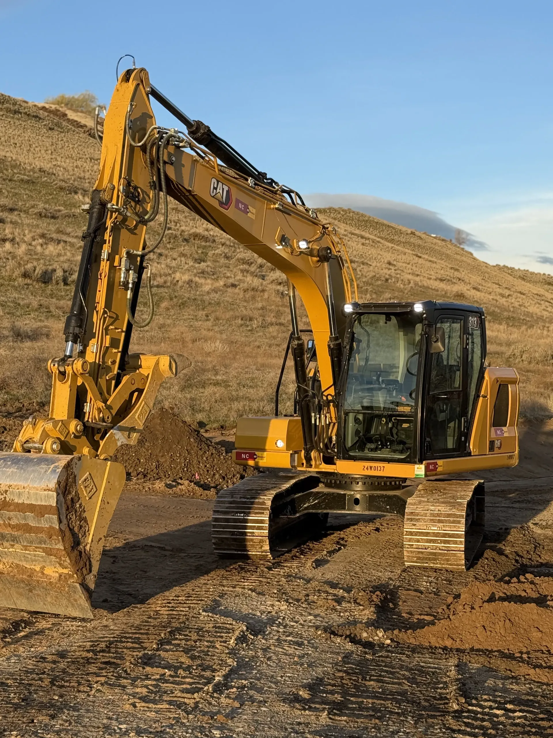 A yellow Caterpillar excavator sits on a dirt construction site against a backdrop of hills under a blue sky.
