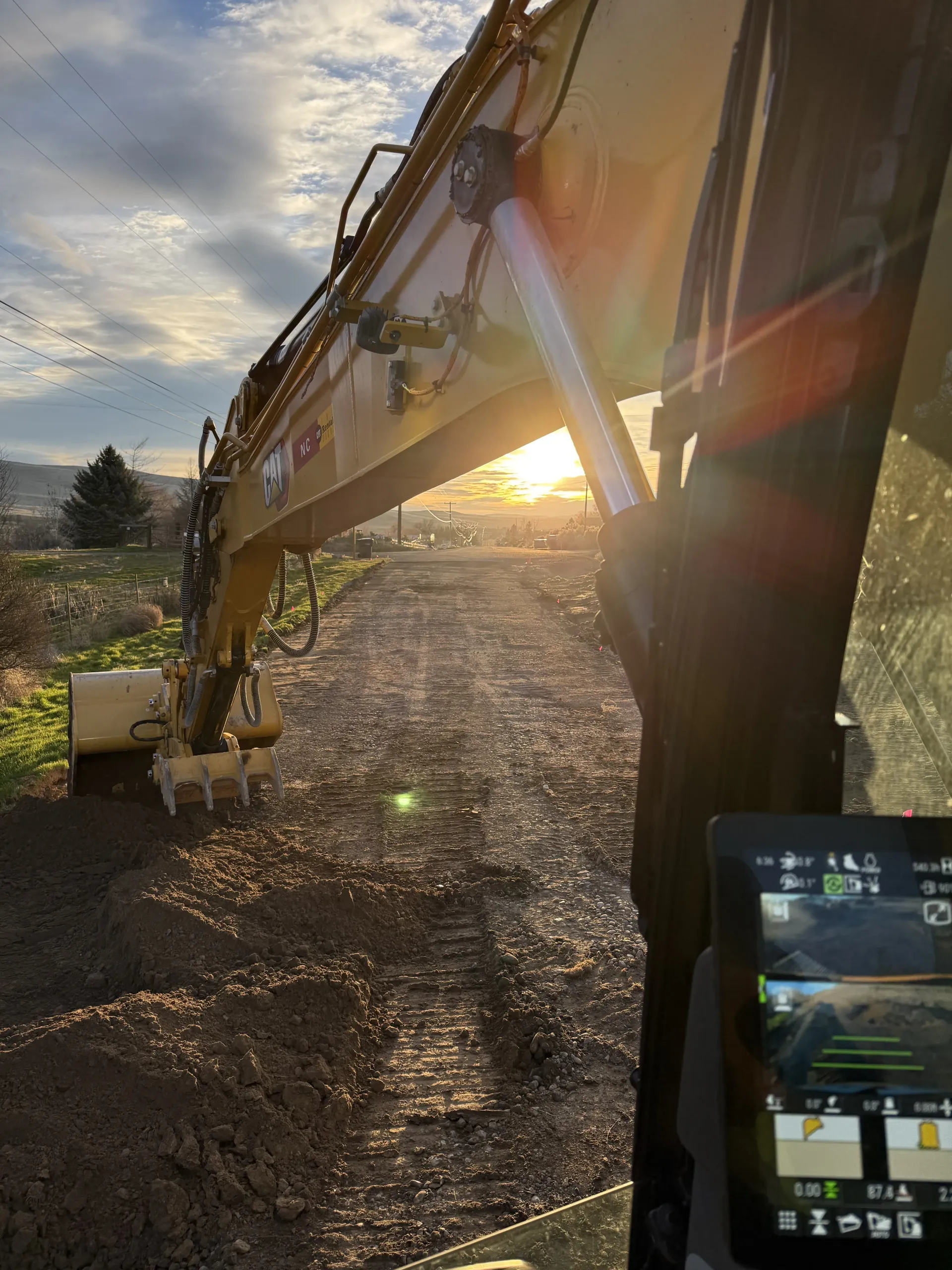 View from inside a yellow excavator cab looking out over a dirt clearing at sunset.