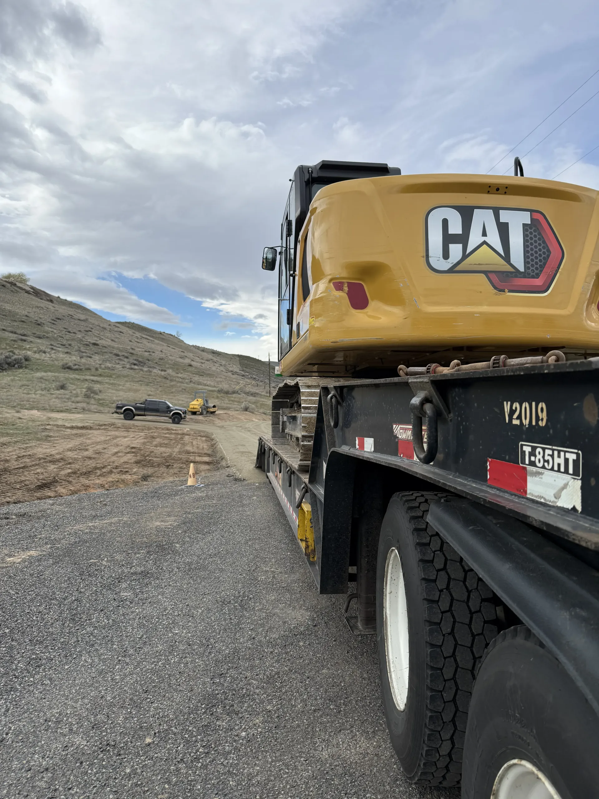A yellow CAT excavator sits on a flatbed trailer on a gravel road against a hilly, rural landscape.