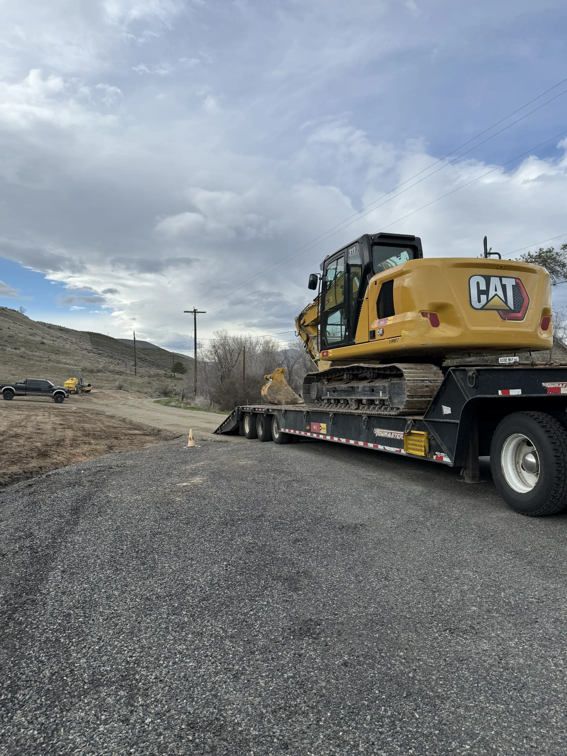 A yellow Caterpillar excavator sits on a flatbed trailer at a gravel-covered construction site under a cloudy sky.
