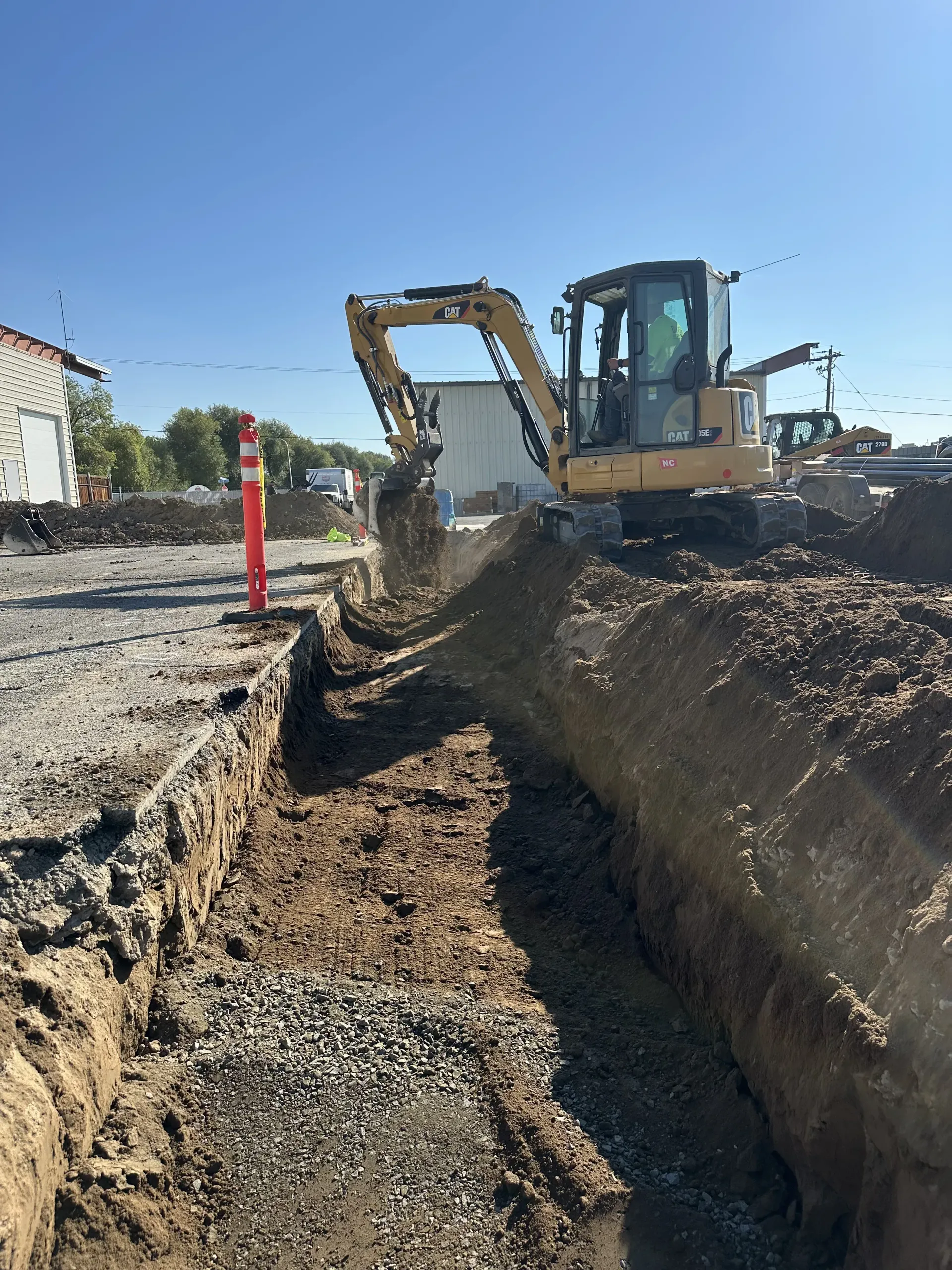 A yellow excavator digs a trench into the dirt ground under a clear blue sky.