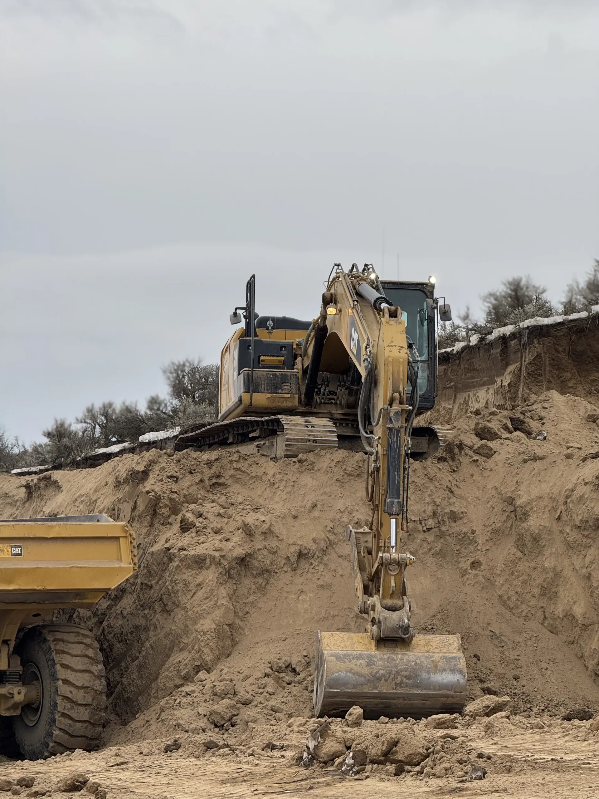 A yellow excavator sits atop a dirt embankment near a construction truck under a cloudy sky.