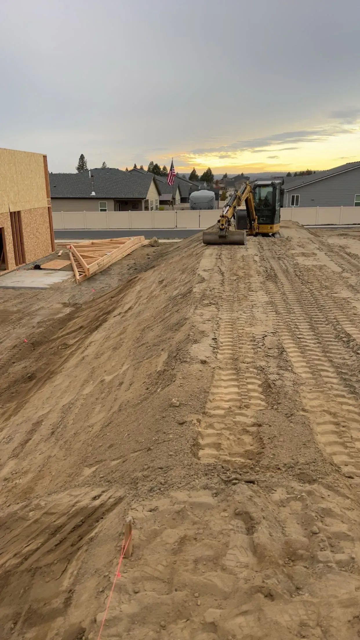 A construction site with a yellow excavator working on a dirt lot near a partially built wooden structure at dusk.