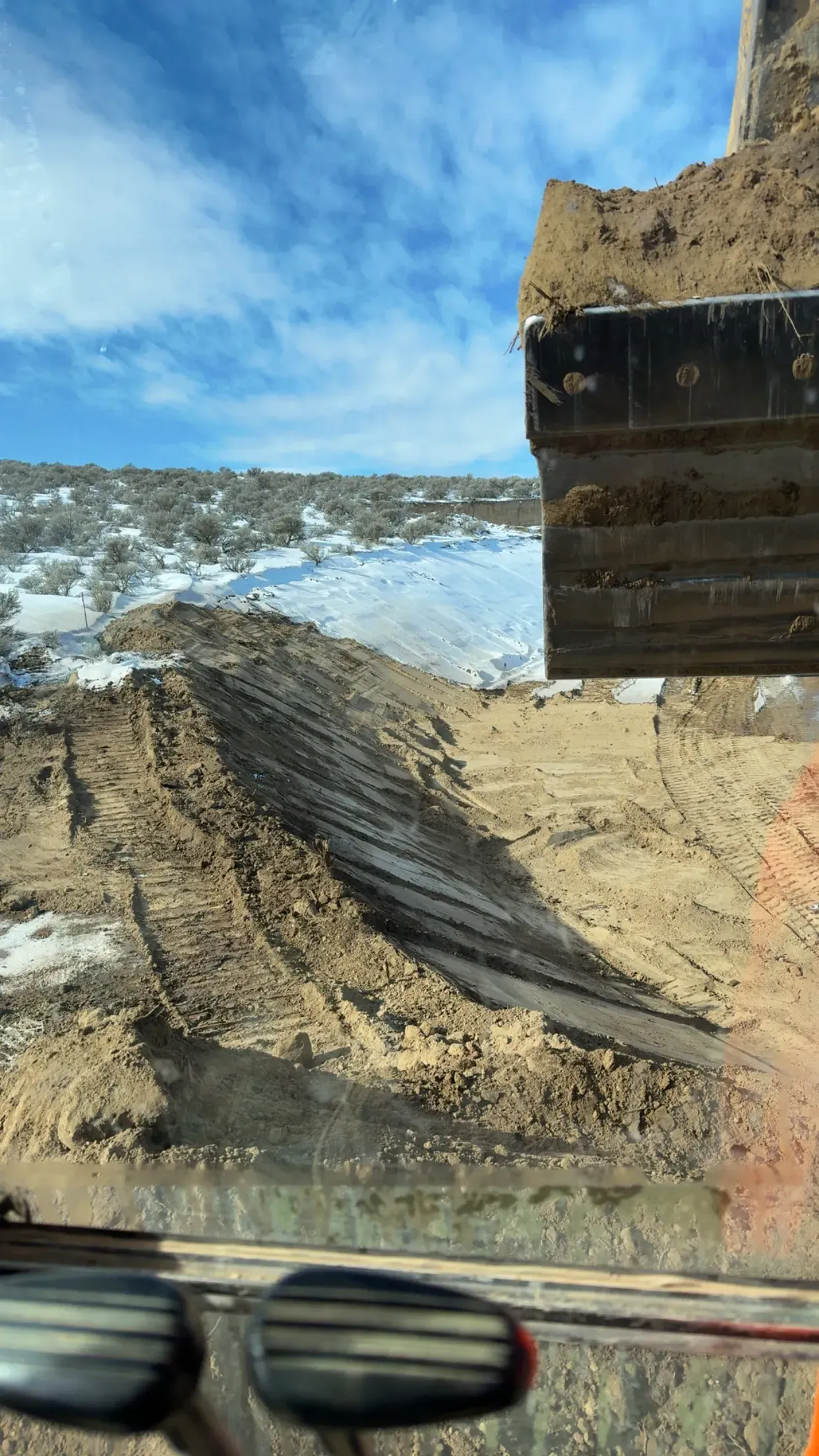 View from an excavator cab looking out over a snow-covered dirt excavation site under a blue, cloudy sky.
