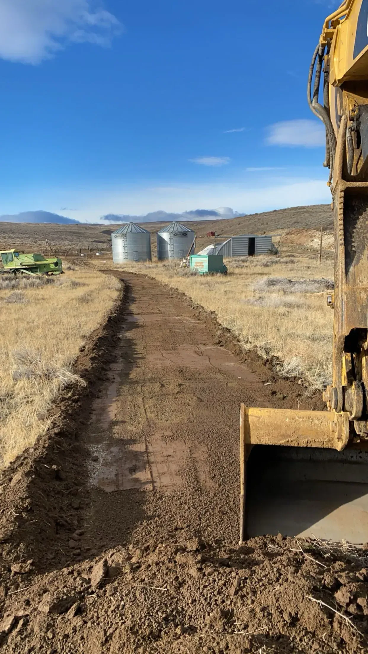 A yellow excavator creates a dirt path leading toward grain silos in a dry, open field under a blue sky.