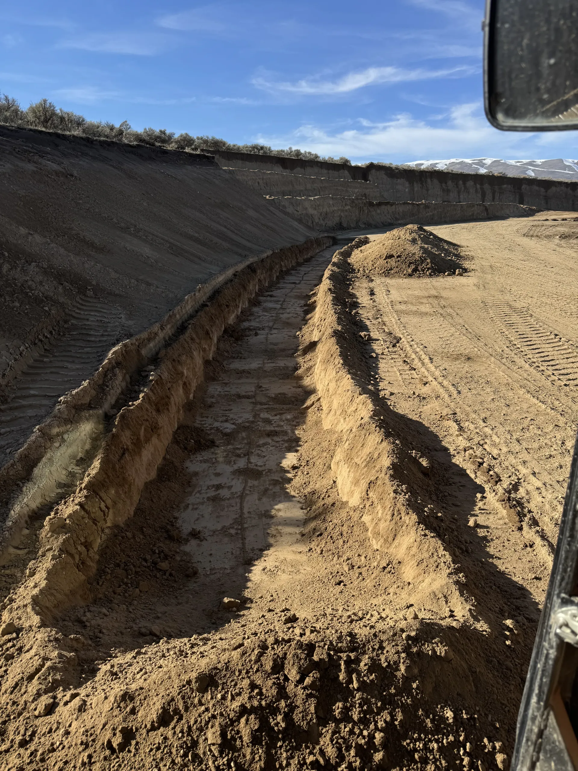 A long, narrow dirt trench curves through a construction site under a clear blue sky, viewed from a vehicle cab.