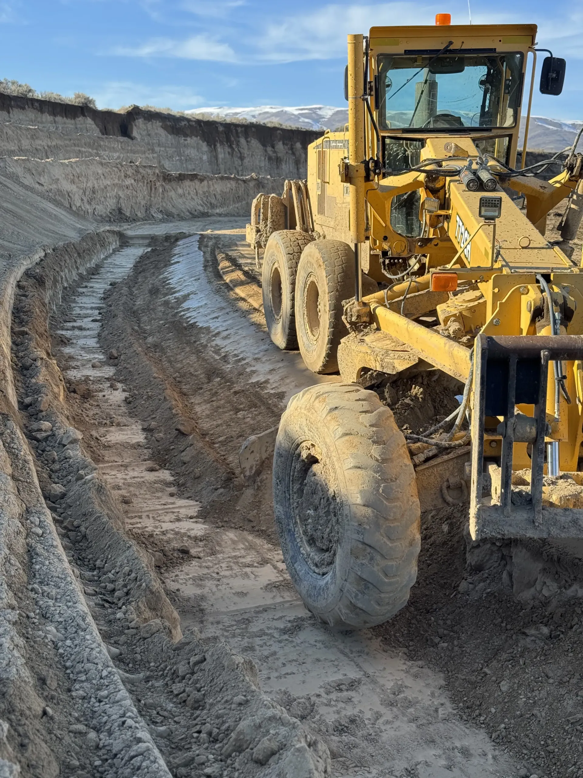 A yellow motor grader working in a muddy construction site near a dirt embankment under a clear blue sky.