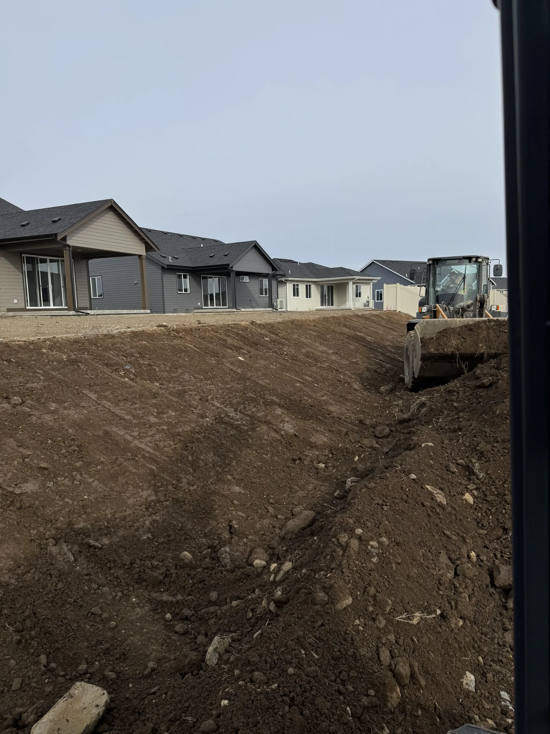 A construction site with a heavy excavator digging a trench in front of several newly built houses under a gray sky.