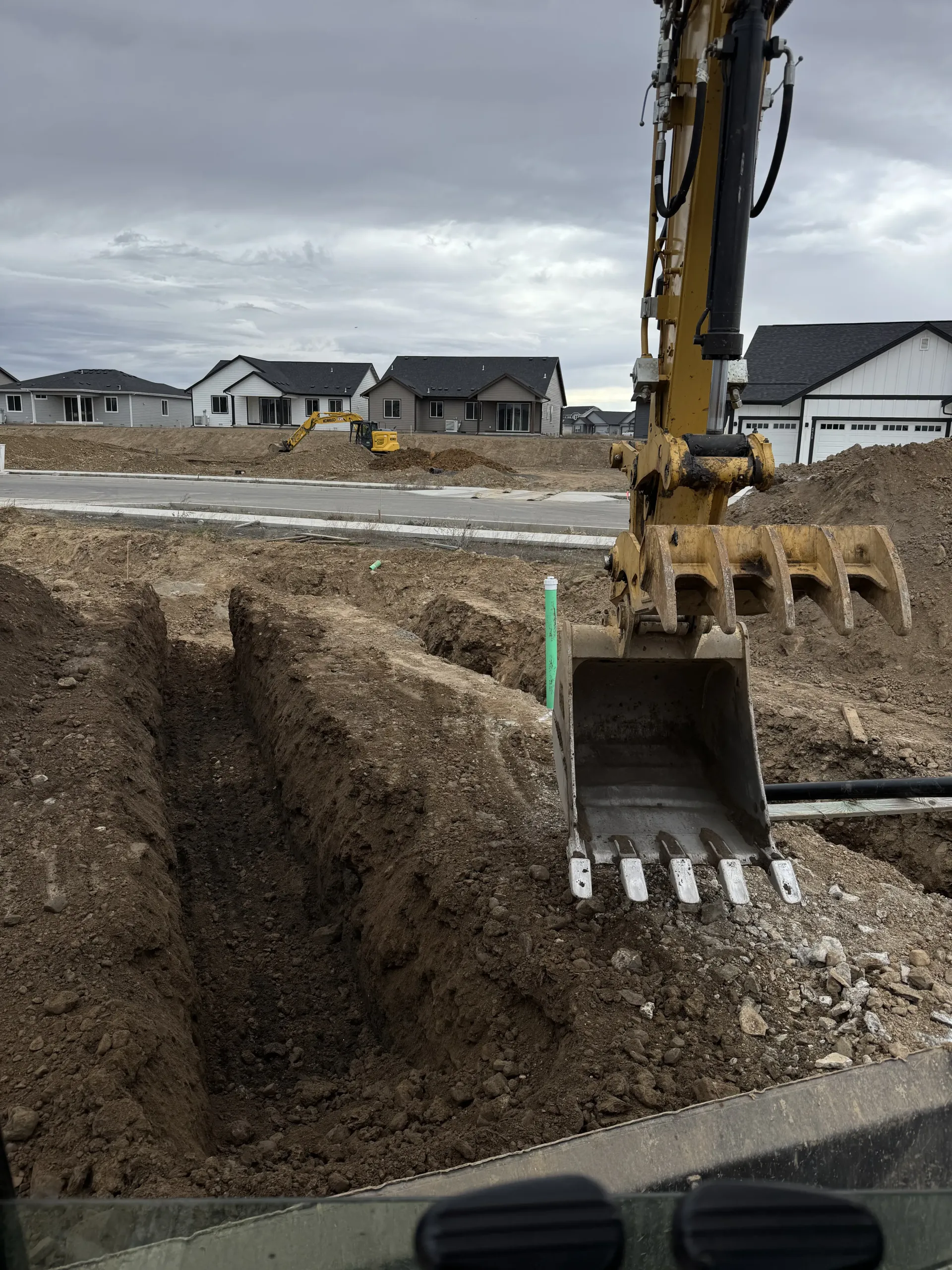 An excavator bucket in the foreground overlooks a freshly dug trench at a suburban construction site.