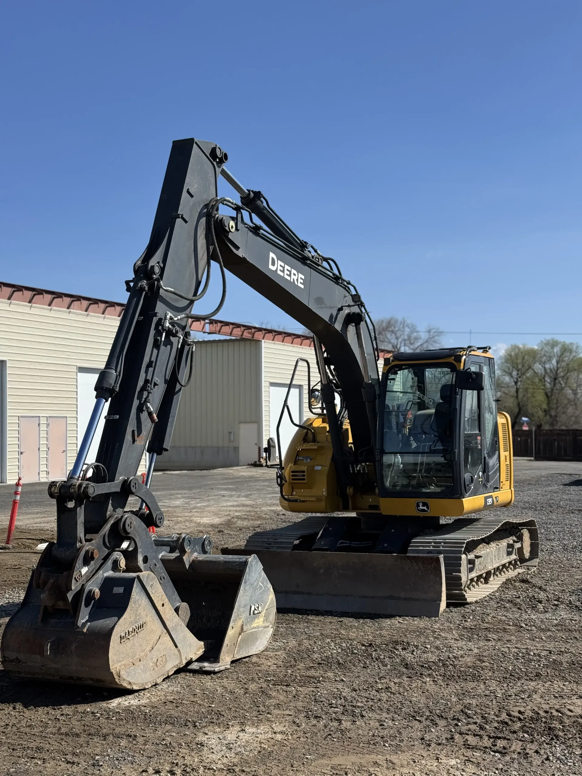 A yellow and gray John Deere excavator parked on a gravel lot in front of a building under a clear blue sky.