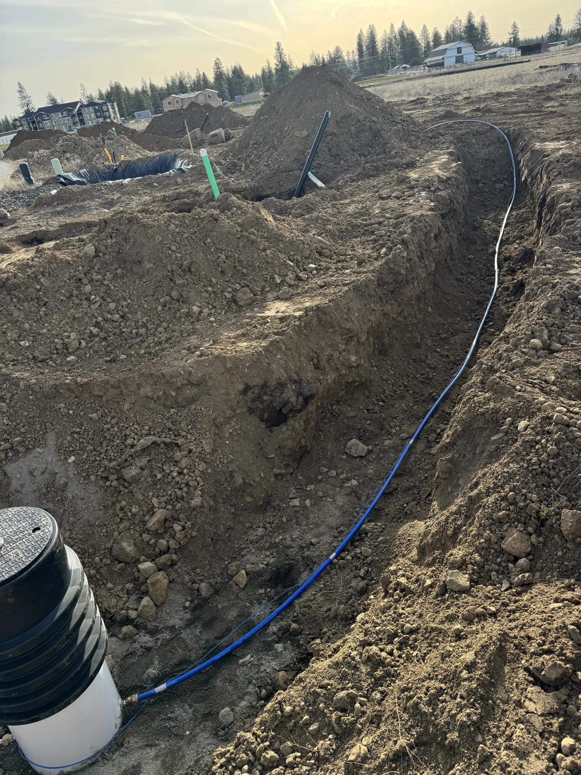 A blue pipe runs through a dirt trench leading to a black utility box at a construction site.