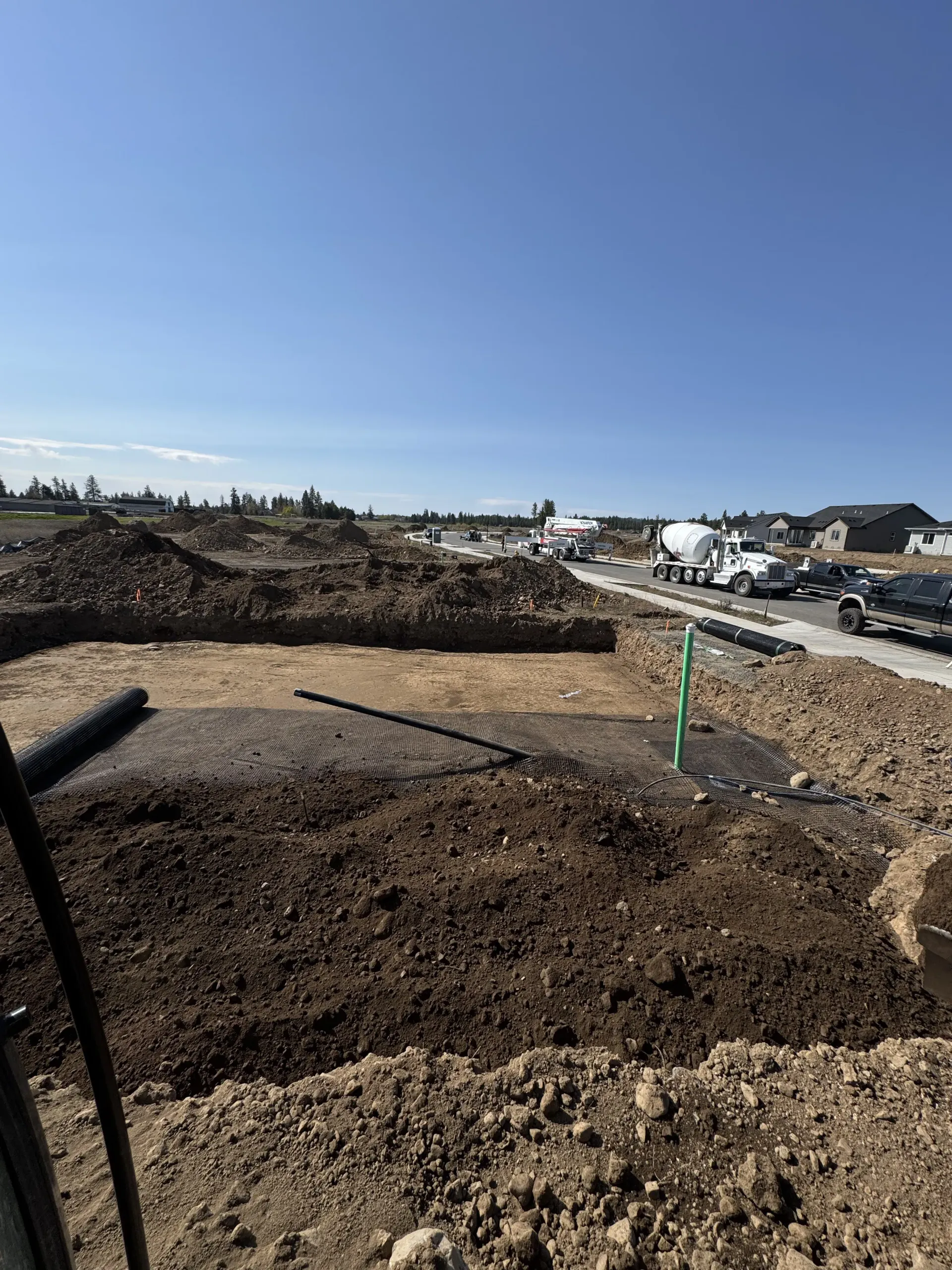 A residential construction site under a clear blue sky, featuring excavated land, mounds of dirt, and parked work vehicles.