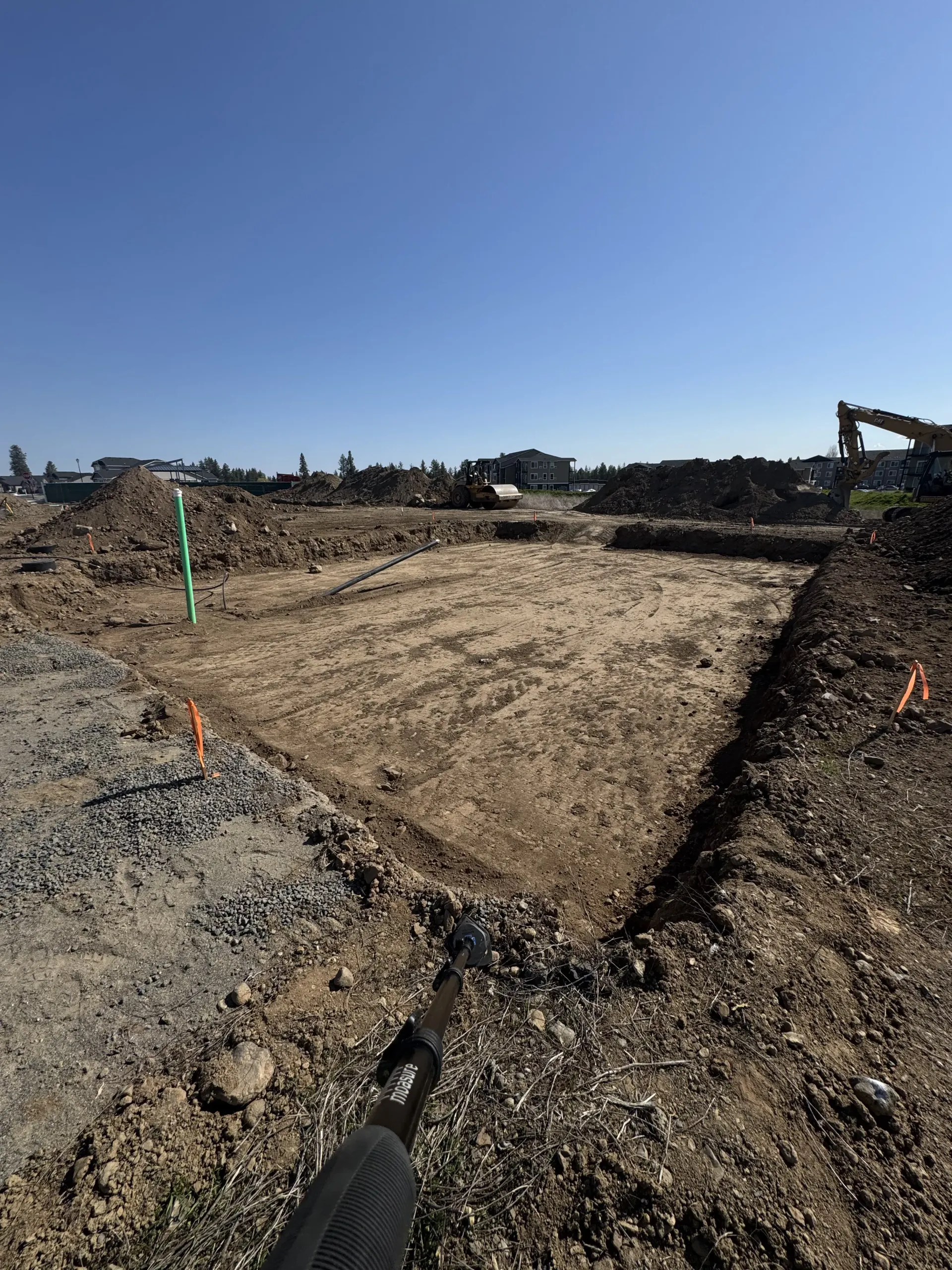 A large rectangular excavation site in a field, with mounds of dirt and construction equipment visible under a blue sky.