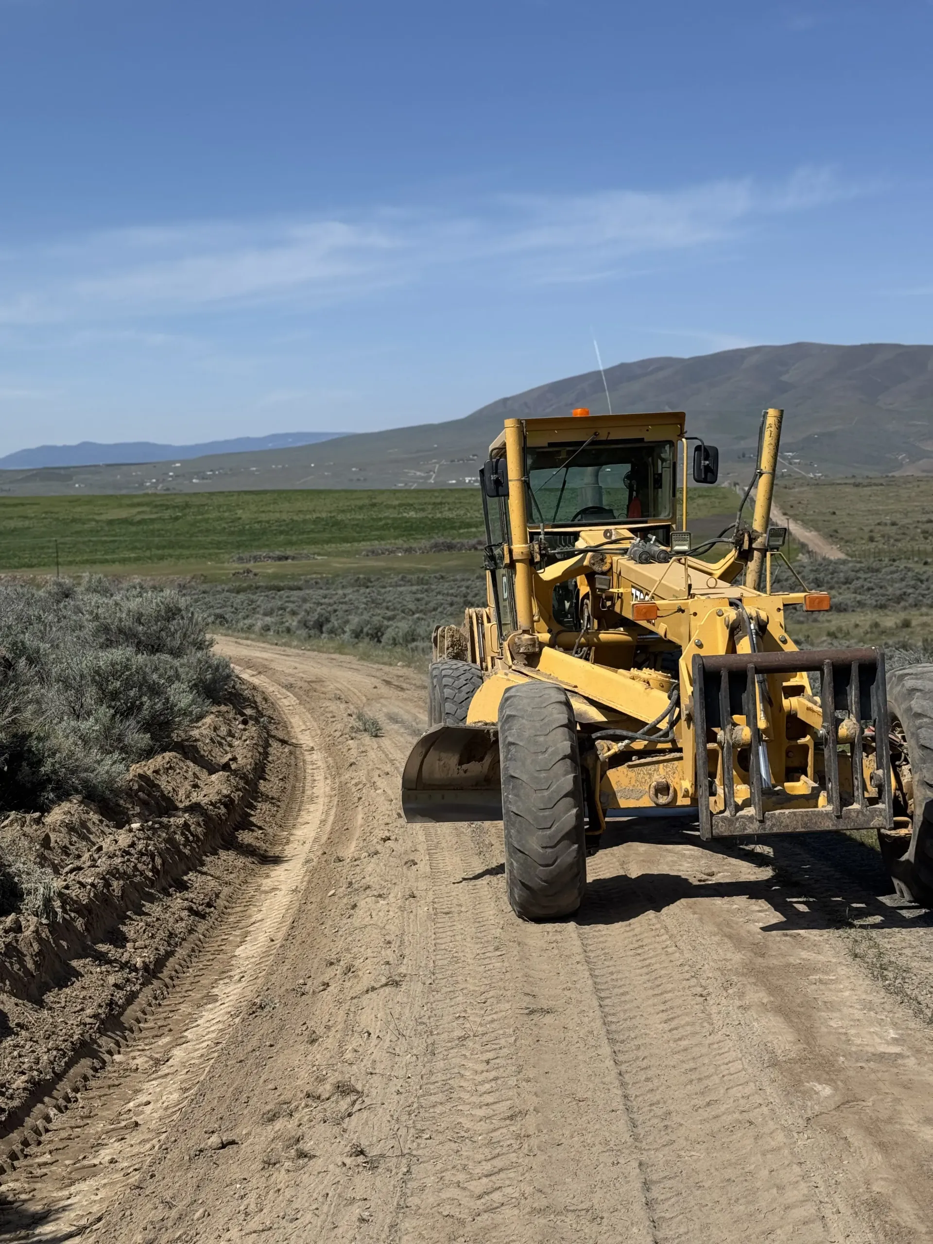 A yellow motor grader drives on a dirt road in a desert landscape with mountains under a blue sky.