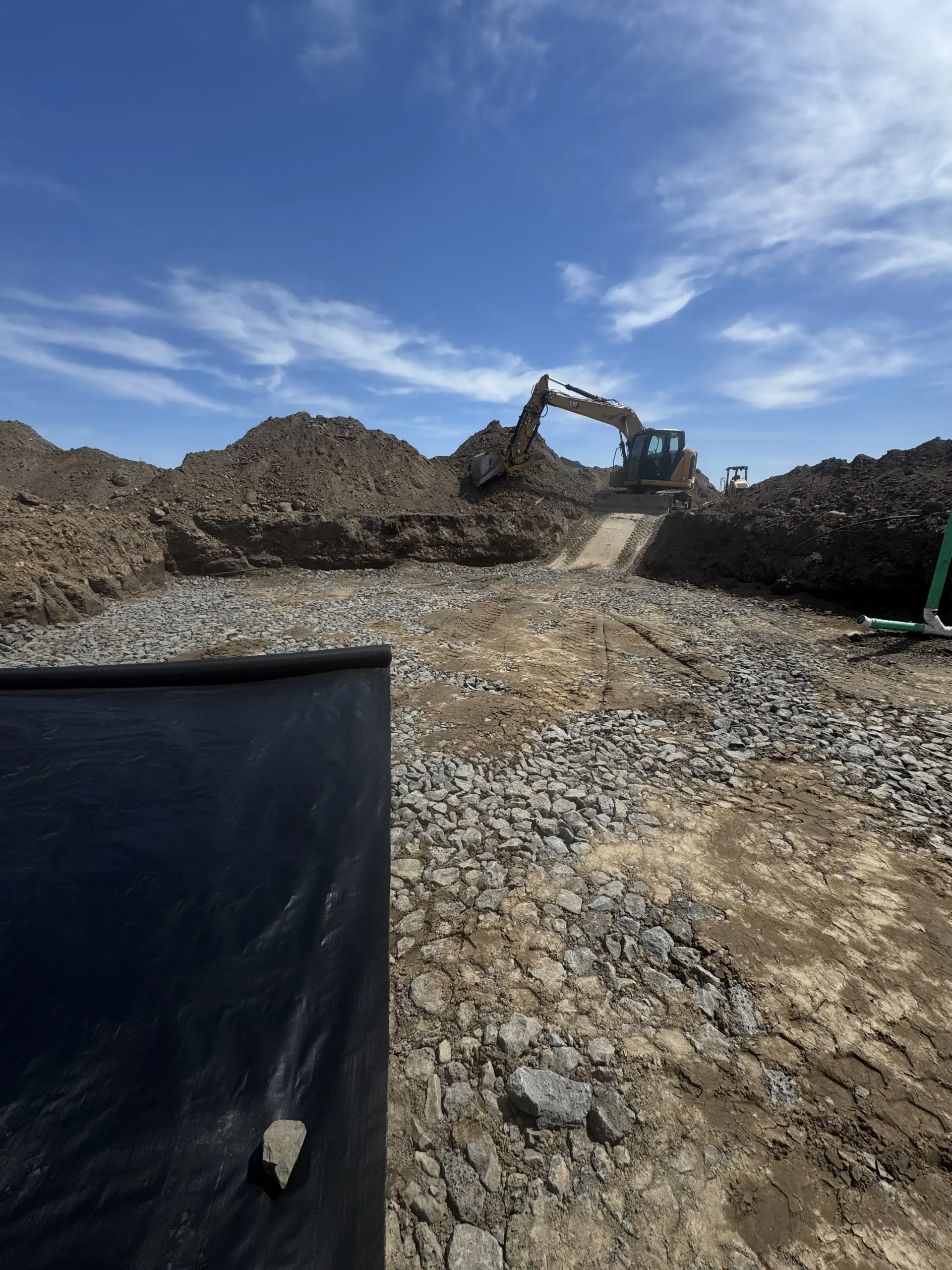 An excavator works on a construction site with piles of excavated dirt under a bright blue sky.